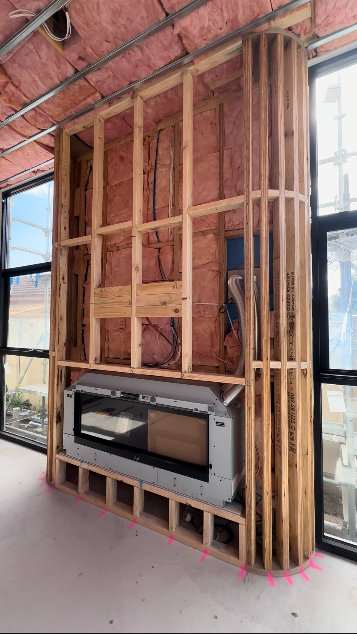 Construction of a wall with an electric fireplace installed beneath a curved corner, with pink insulation and exposed wooden framing, in a room with large windows.