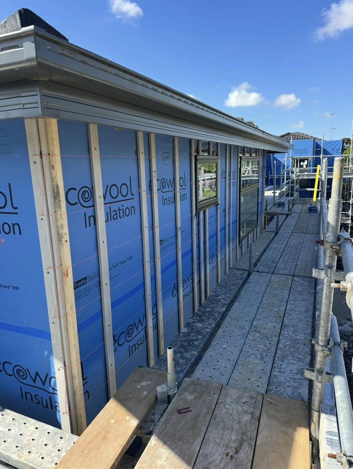 Construction site with a building under construction, blue insulation panels, scaffolding, and a partly finished window, under a clear blue sky.