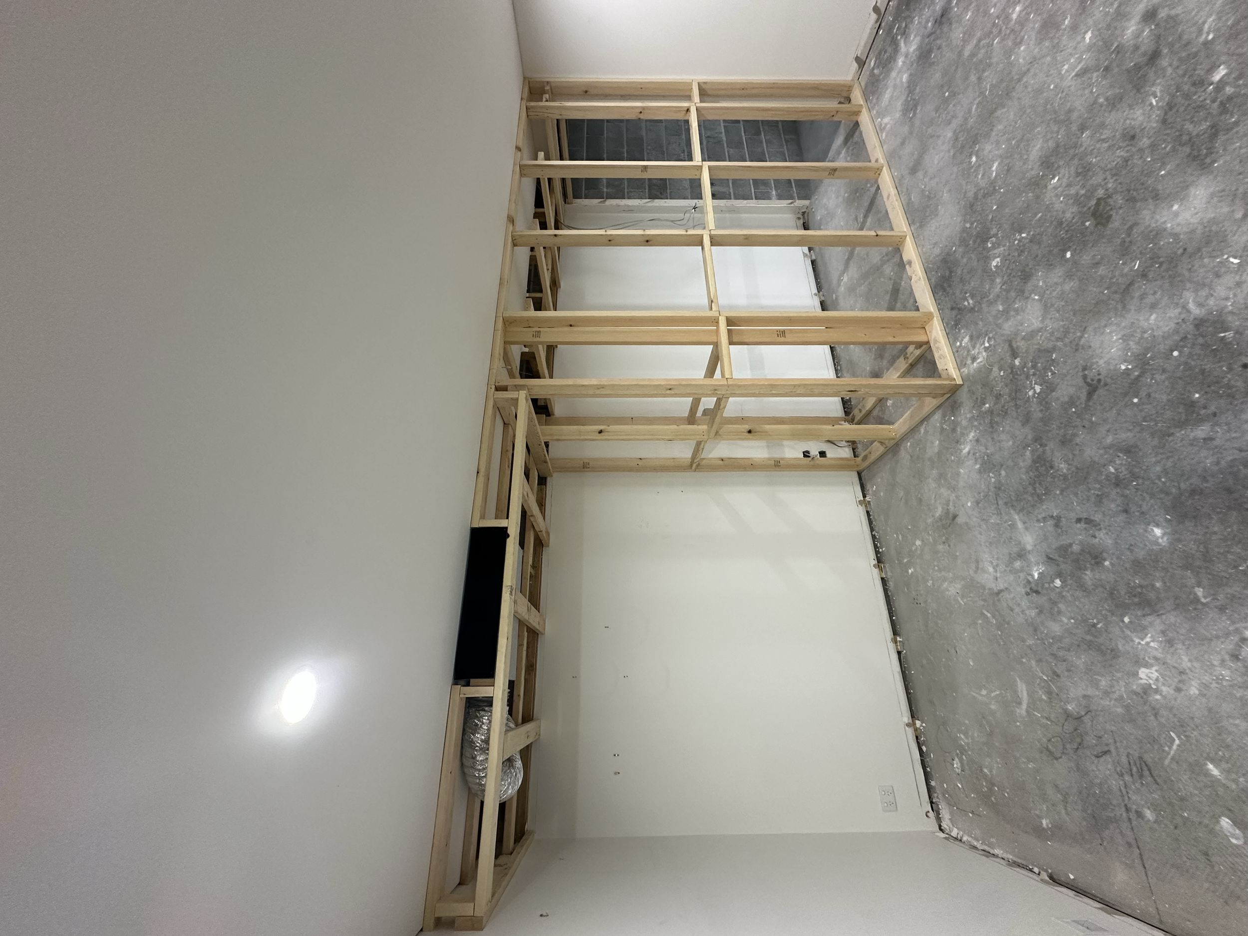 An unfinished room with a wooden frame structure, concrete flooring, and a white wall with an electrical outlet.