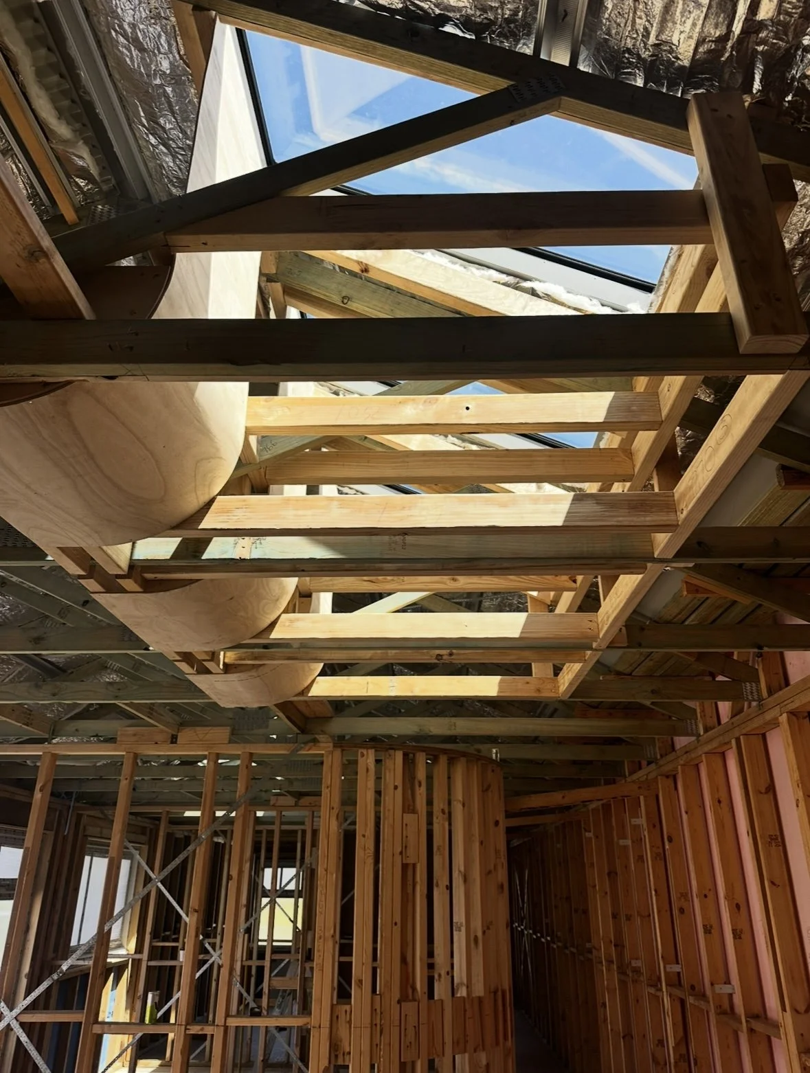 Interior view of a house under construction showing wooden framing, large window openings, and a blue sky outside.