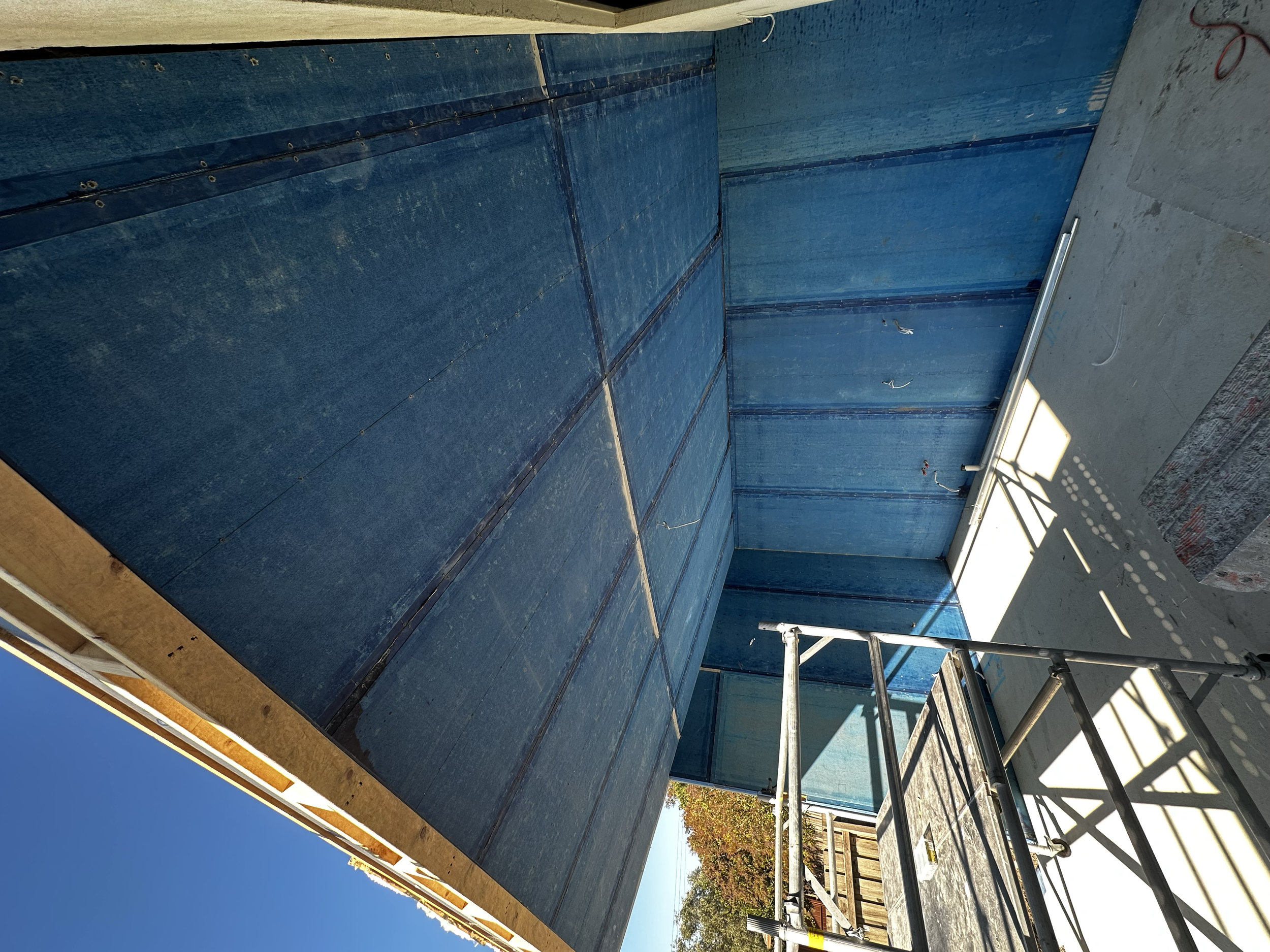 Construction site with blue wall panels, scaffolding, and sunlight casting shadows, under a clear blue sky.