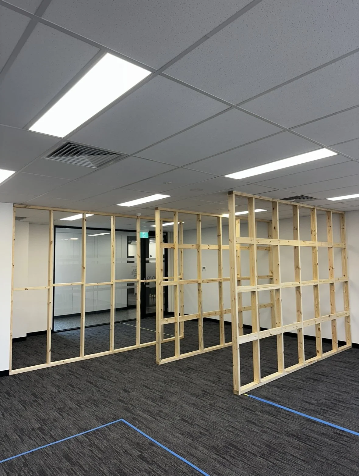 A partially constructed wooden partition wall in an office with gray carpet, ceiling lights, and a glass door in the background.