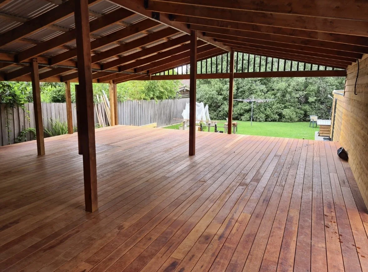 Empty wooden patio with a roof, overlooking a backyard with green grass, trees, and a clothesline.