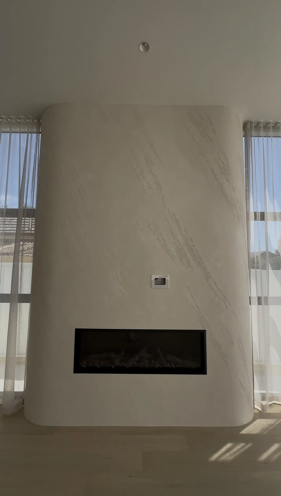 Modern interior fireplace with textured white wall, two large windows with sheer curtains, and natural light.