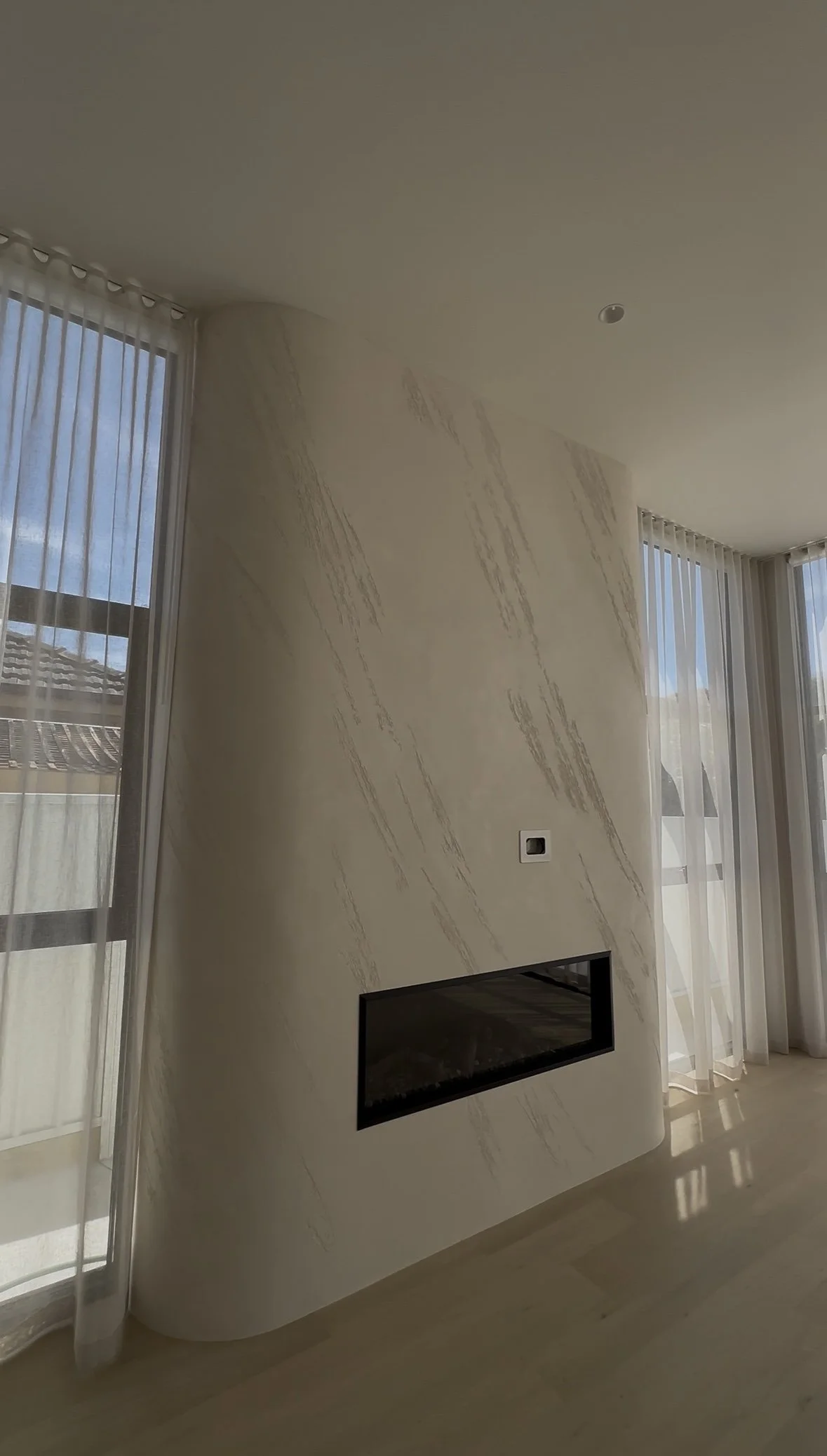 A modern interior living room with floor-to-ceiling windows, sheer white curtains, a rounded white wall with marble vein accents, and a rectangular fireplace built into the wall.