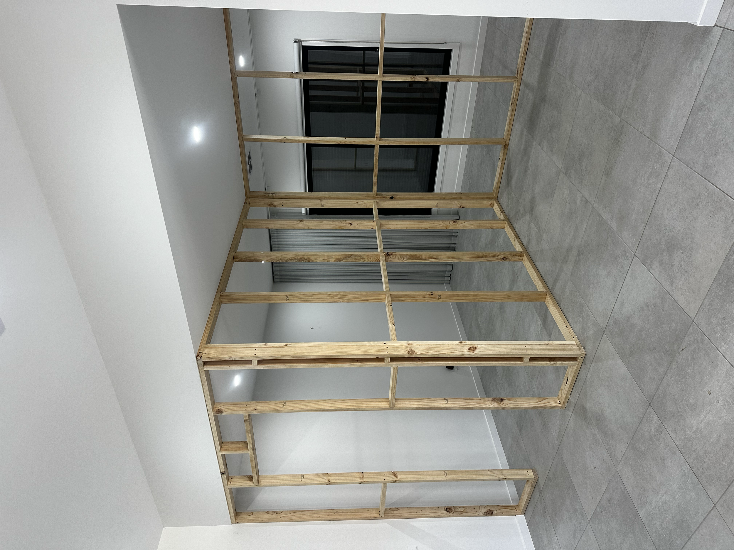 Unfinished wooden wall frame in a room with gray tile flooring, white walls, and a large sliding glass door.