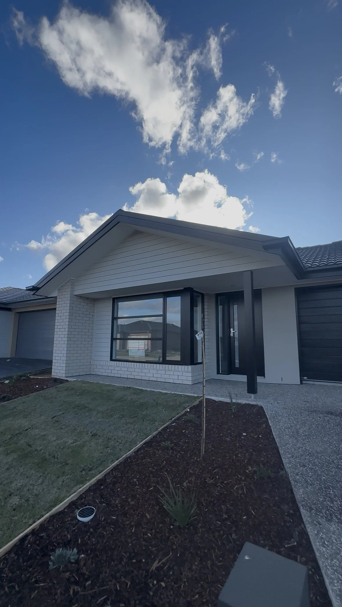 New house with white brick and black window frames, front yard with small garden, driveway, and blue sky with clouds.