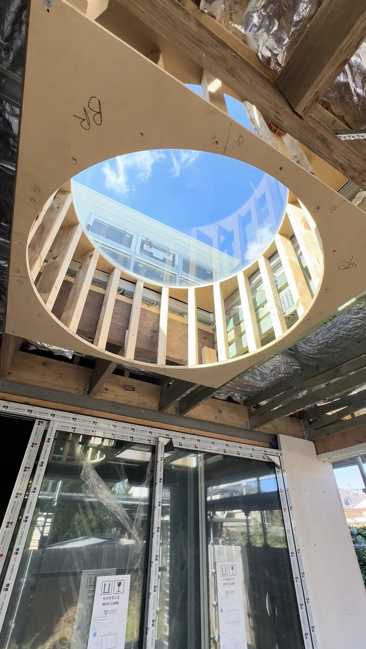 Construction site with a circular opening in the ceiling for a skylight, showing blue sky and clouds above. There are wooden beams and construction materials around, and windows with protective stickers labeled 'Handle with Care' on the lower part of