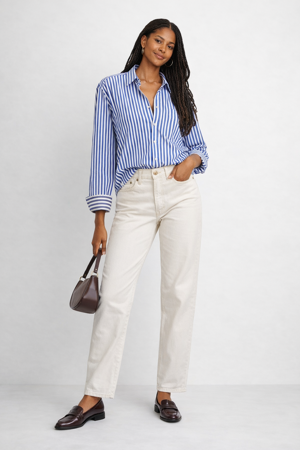 Woman wearing cream jeans with a blue striped button-down shirt and brown loafers styled with a shoulder bag