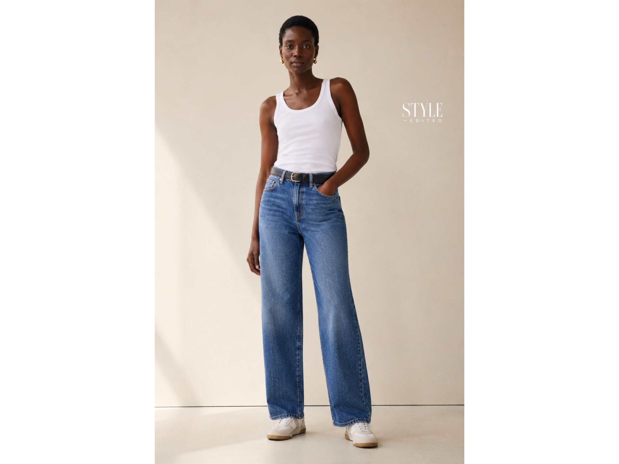 Dark-skinned woman wearing a fitted white tank with relaxed blue jeans and low-profile sneakers in a warm neutral studio.