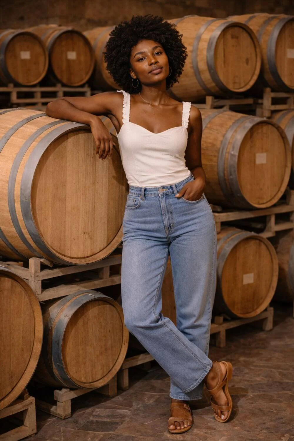 Woman wearing light wash jeans and a white camisole styled for a casual winery outfit inside a wine cellar.