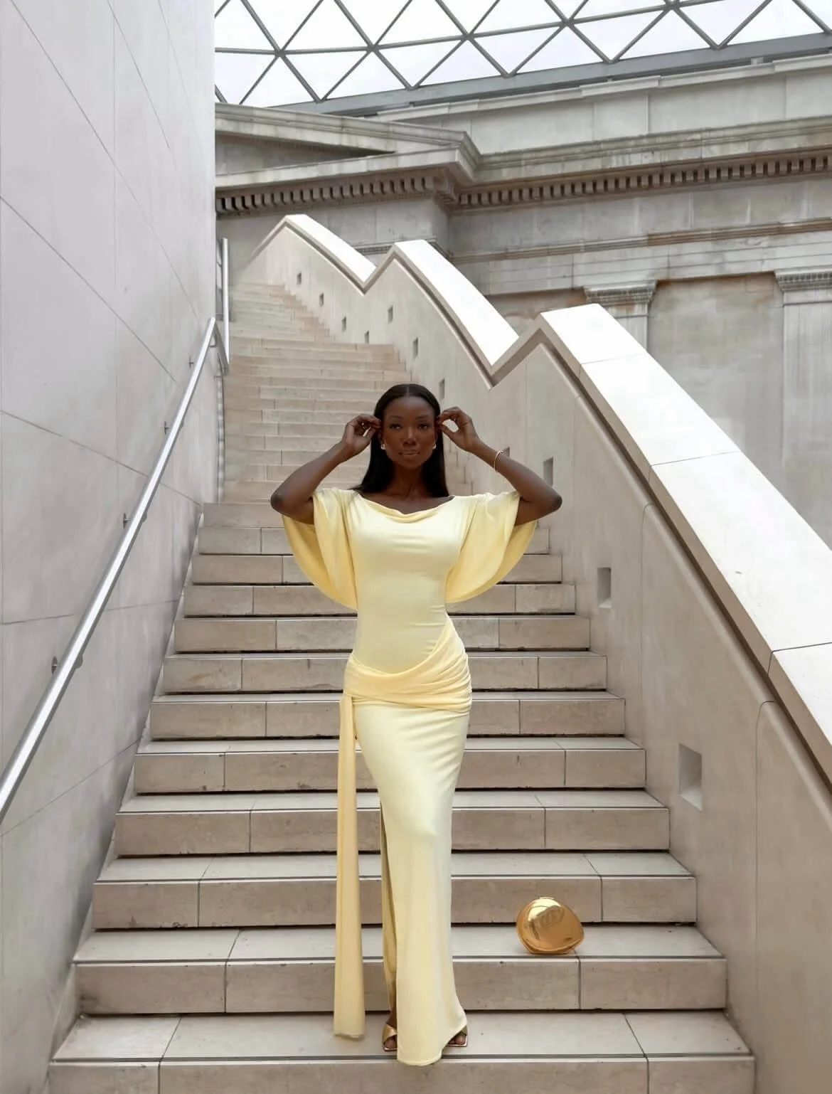 Sia wearing a yellow draped midi dress with soft sleeves and waist gathering, standing on stairs showing a defined silhouette and structured fit