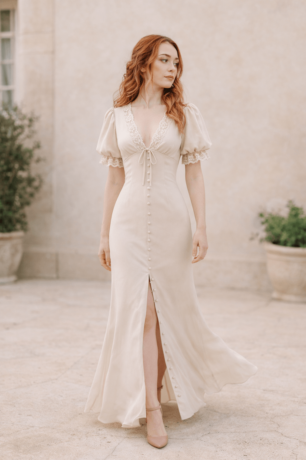 Bride wearing a romantic vintage-inspired lace elopement dress with puff sleeves and front buttons, walking in a light neutral garden villa setting with dusty rose heels