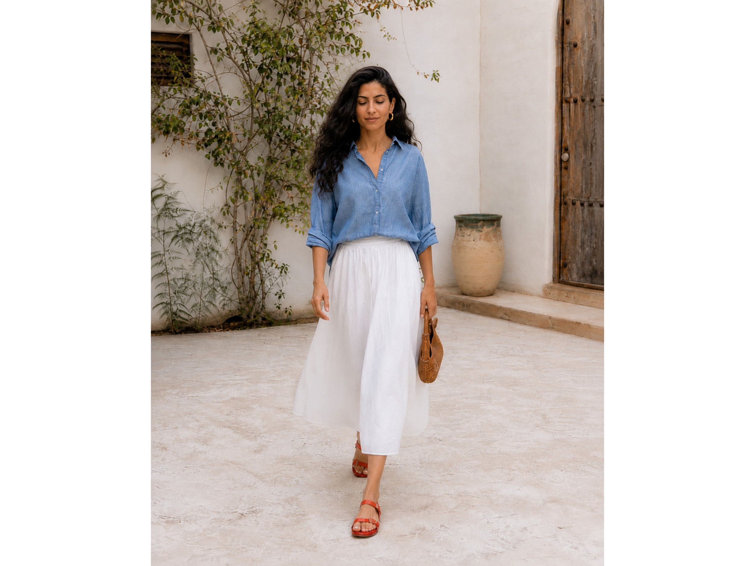 Middle Eastern woman wearing a chambray button-down shirt, white flowy midi skirt, red sandals, and a woven bag while walking through an open-air courtyard.