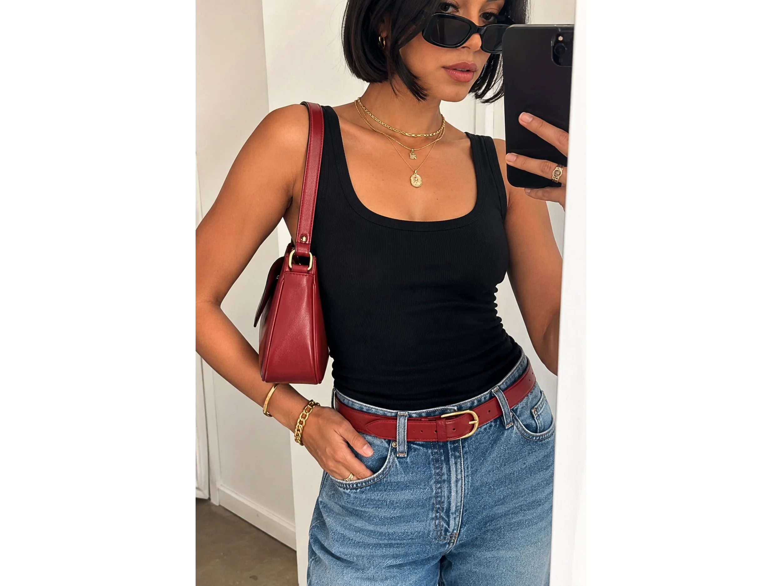 Latina woman in a black tank top and relaxed blue jeans styled with a red belt, red shoulder bag, and layered gold jewelry.