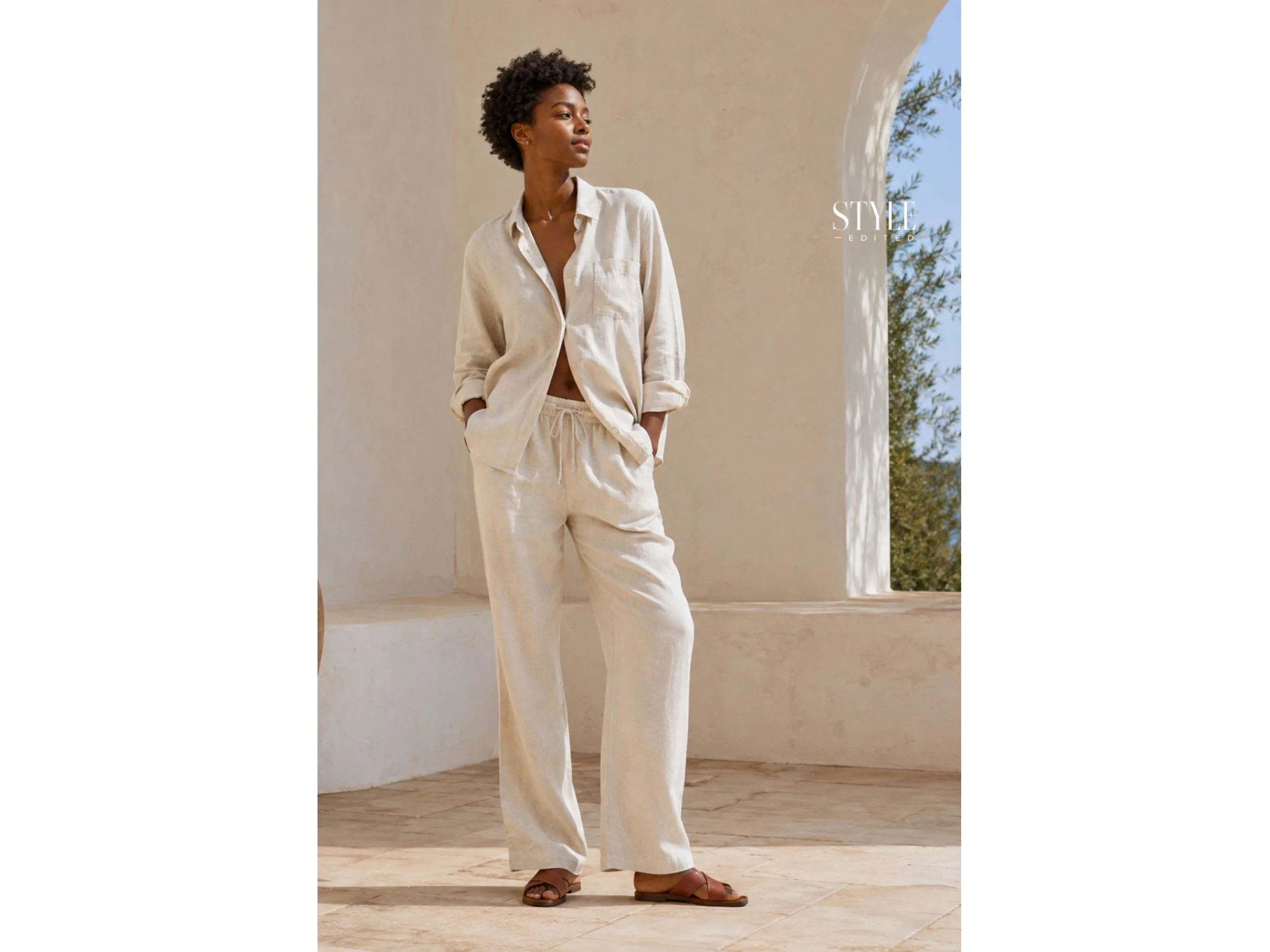 Black woman wearing a relaxed cream linen matching set with brown leather sandals on a sunlit Mediterranean-style terrace.