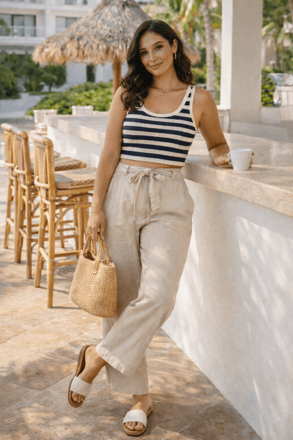 Woman wearing striped cropped tank top with high-waisted linen pants holding woven handbag at outdoor resort bar