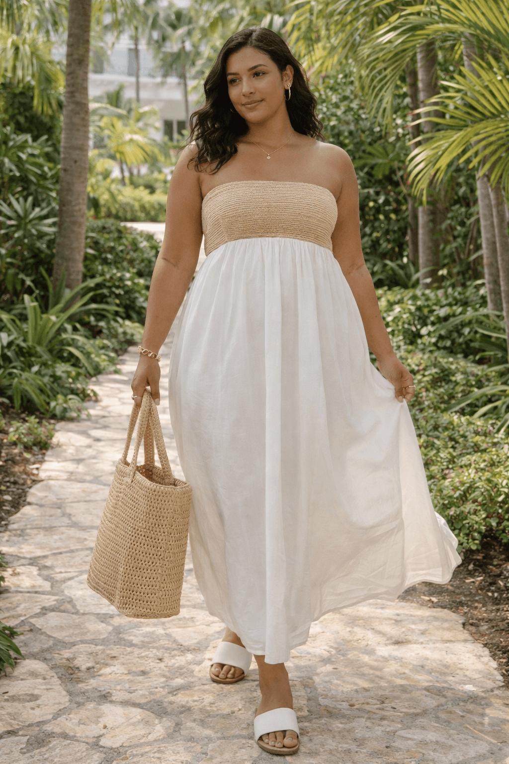 Woman wearing strapless neutral maxi dress with woven tote and slide sandals walking along tropical resort garden path