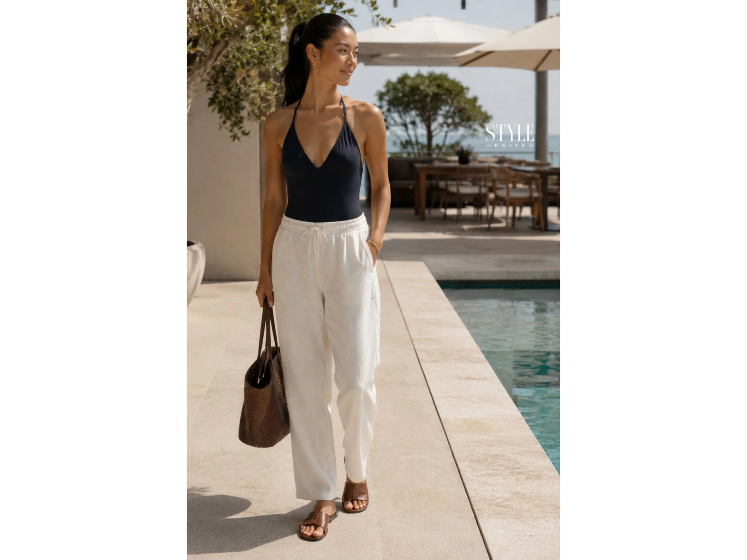 East Asian woman wearing a dark one-piece swimsuit with loose white pull-on pants and leather slides by a pool.