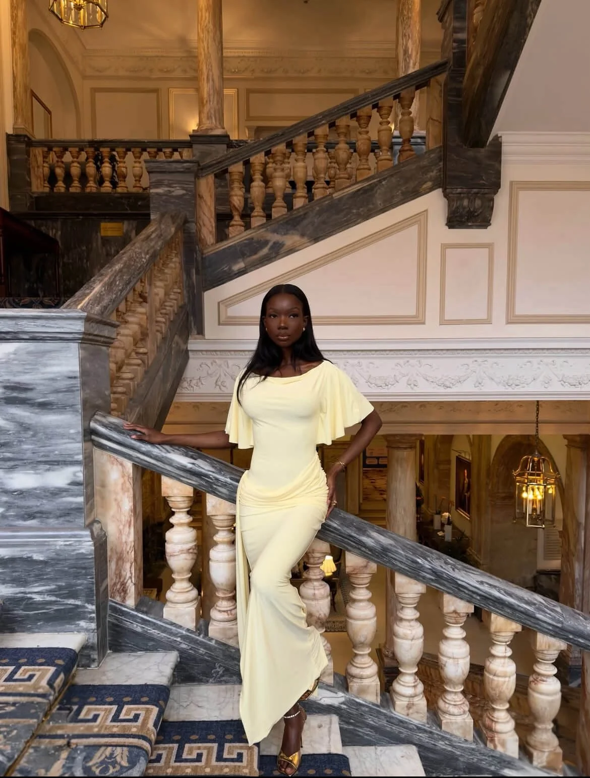 Sia wearing a yellow draped midi dress with a defined waist and soft structure, standing on stairs in an elegant interior