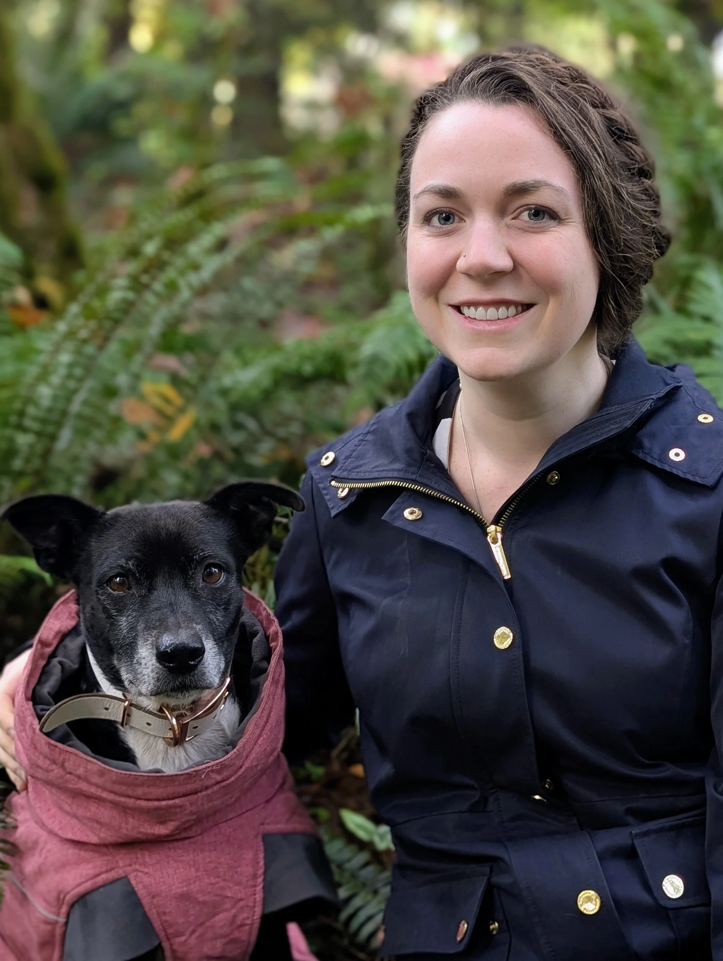A woman with short brown hair wearing a navy blue jacket, smiling, outdoors with greenery, next to a black and white dog in a pink jacket with a collar.