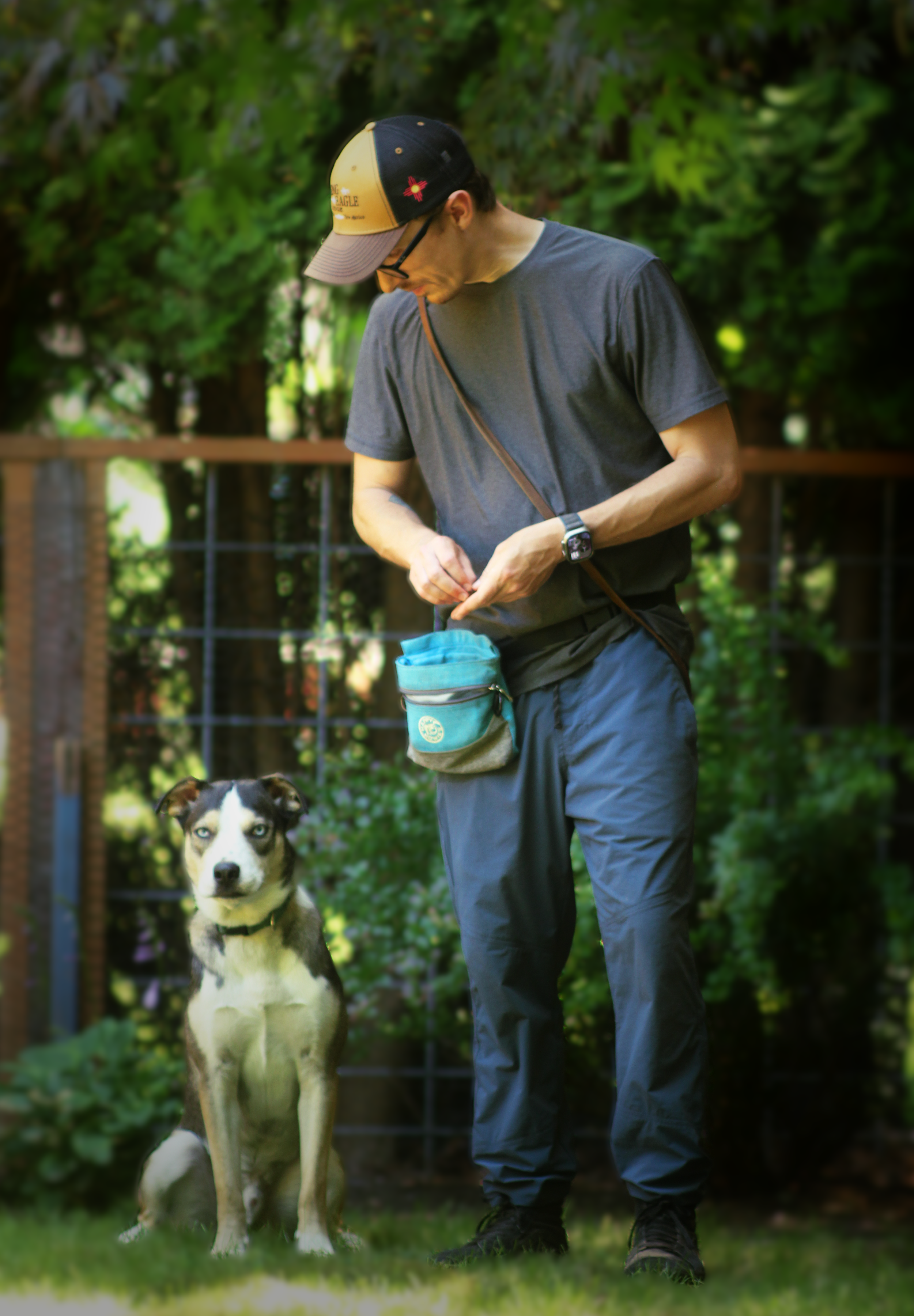 A man wearing a grey shirt and blue pants stands looking down at a white and grey dog with light blue eyes sitting next to the man.