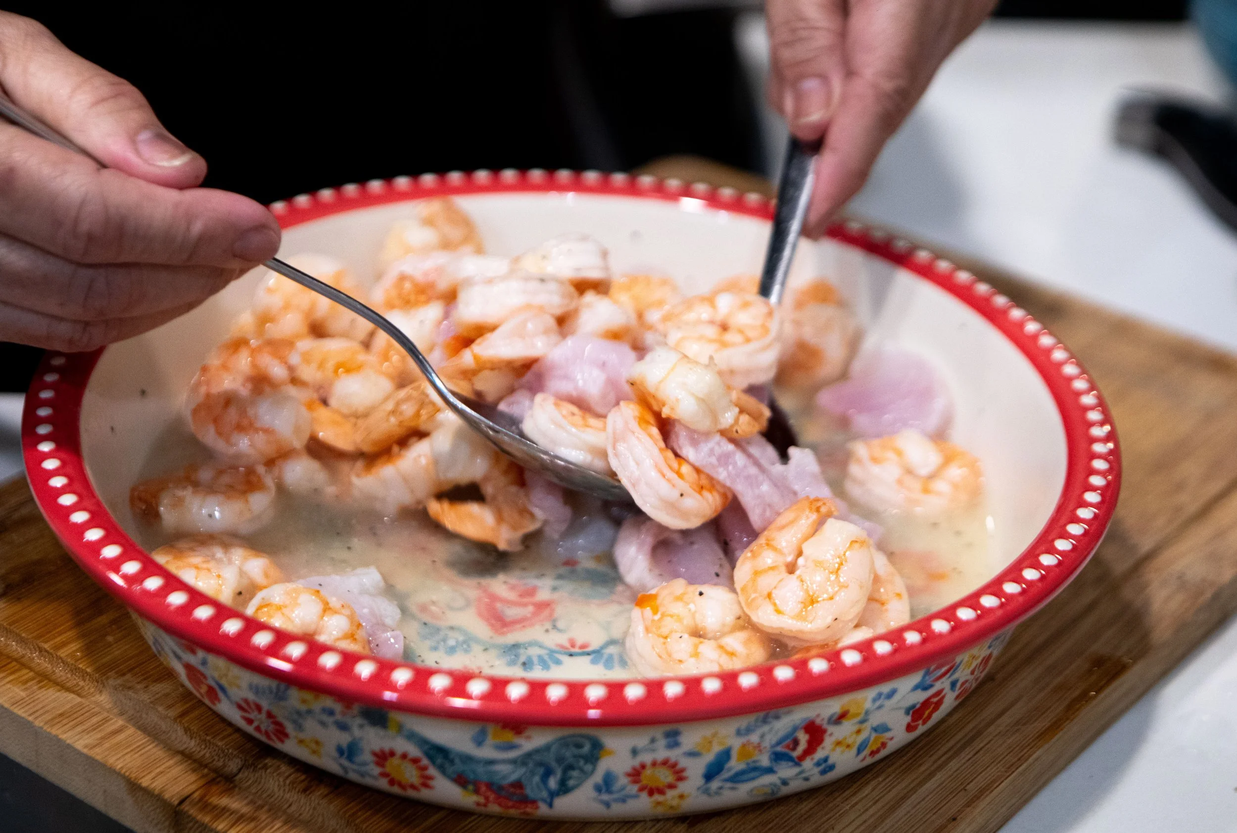 Hands mixing shrimp and sliced ham in a decorative ceramic bowl with a creamy sauce on a wooden surface.