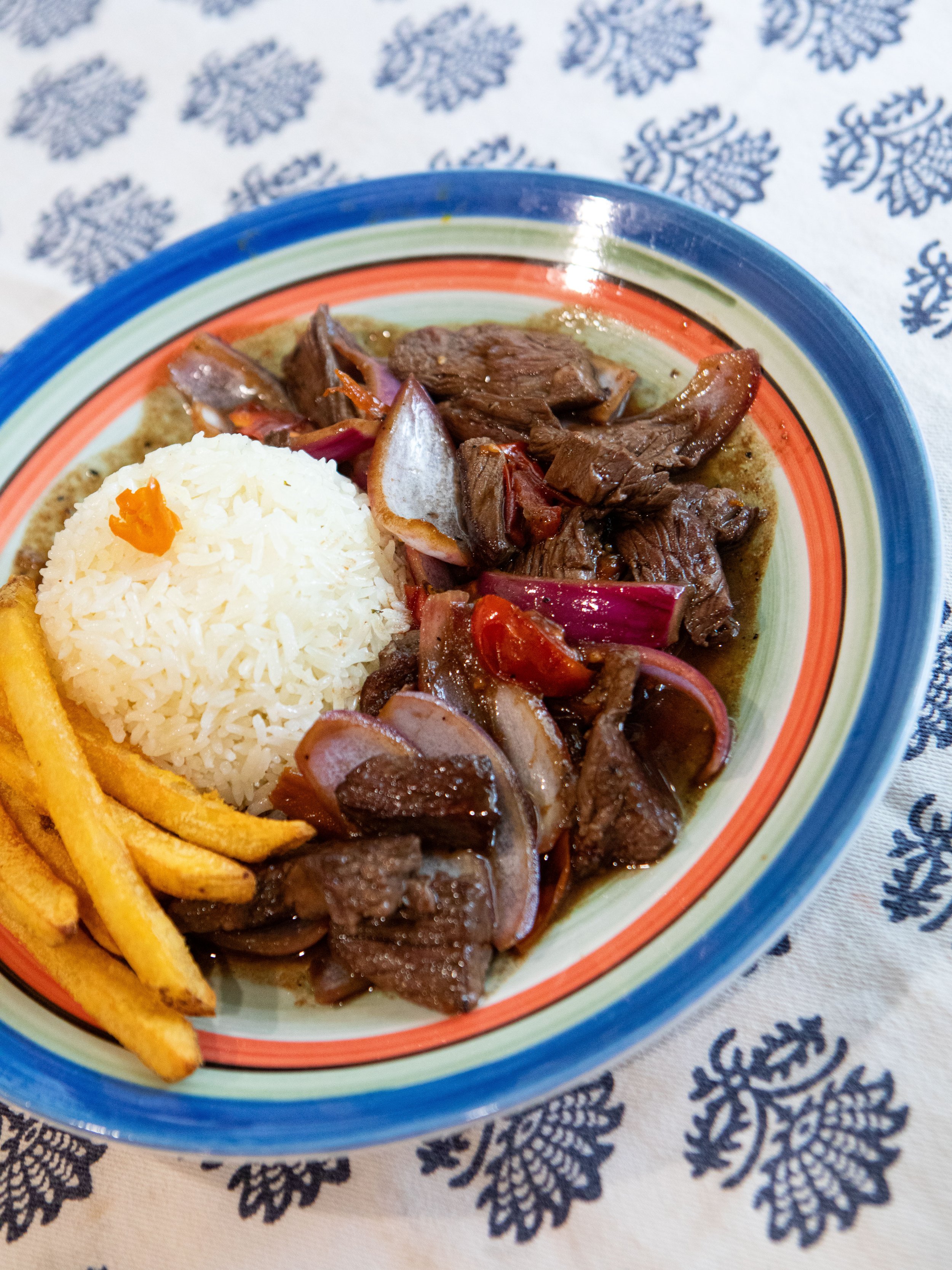 A colorful bowl of food containing rice, beef stew with onions and cherry tomatoes, and French fries on a patterned tablecloth.