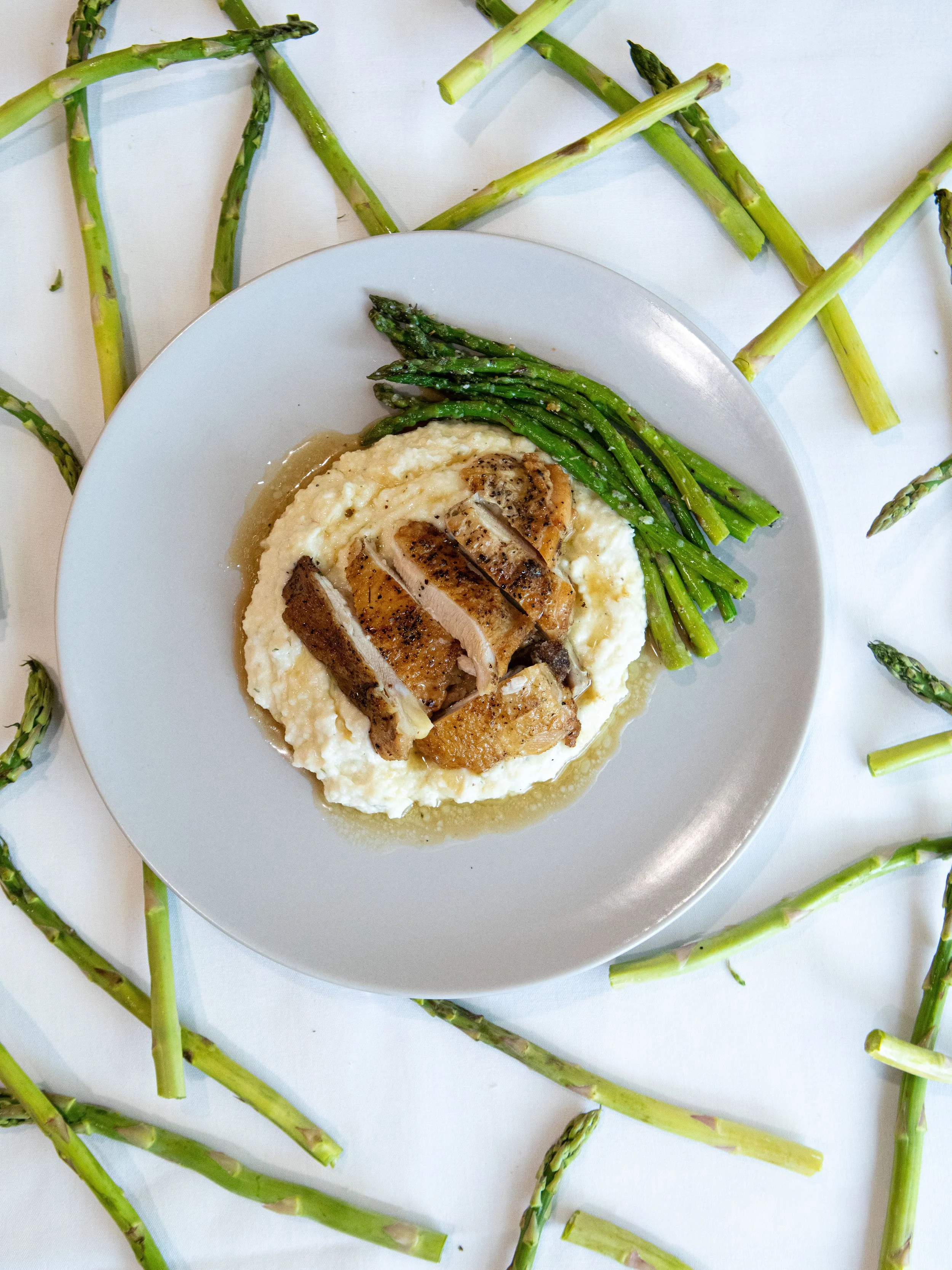 Plate of cooked asparagus, mashed potatoes, and sliced roasted chicken on a white plate, surrounded by raw asparagus spears on a white surface.