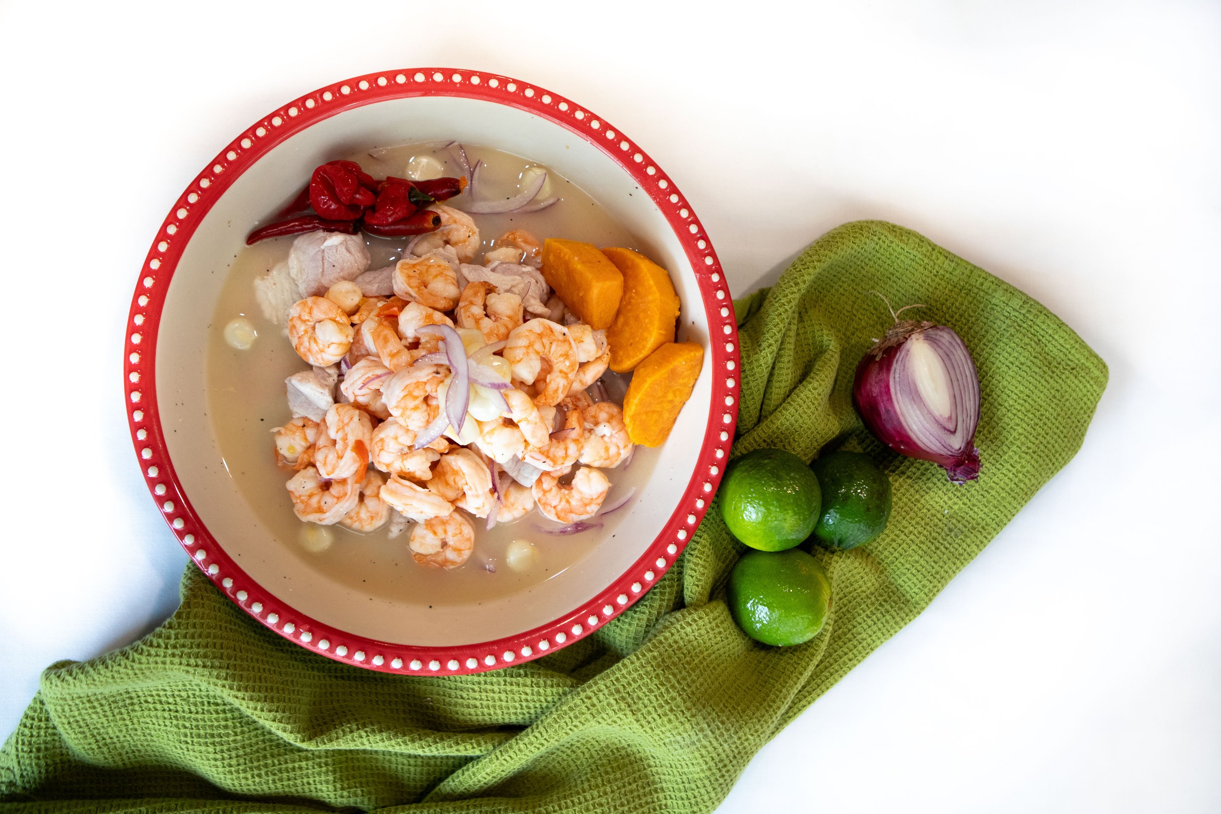 A bowl of Mexican shrimp cocktail with lime wedges, red chili peppers, and a halved red onion on a lime green cloth.