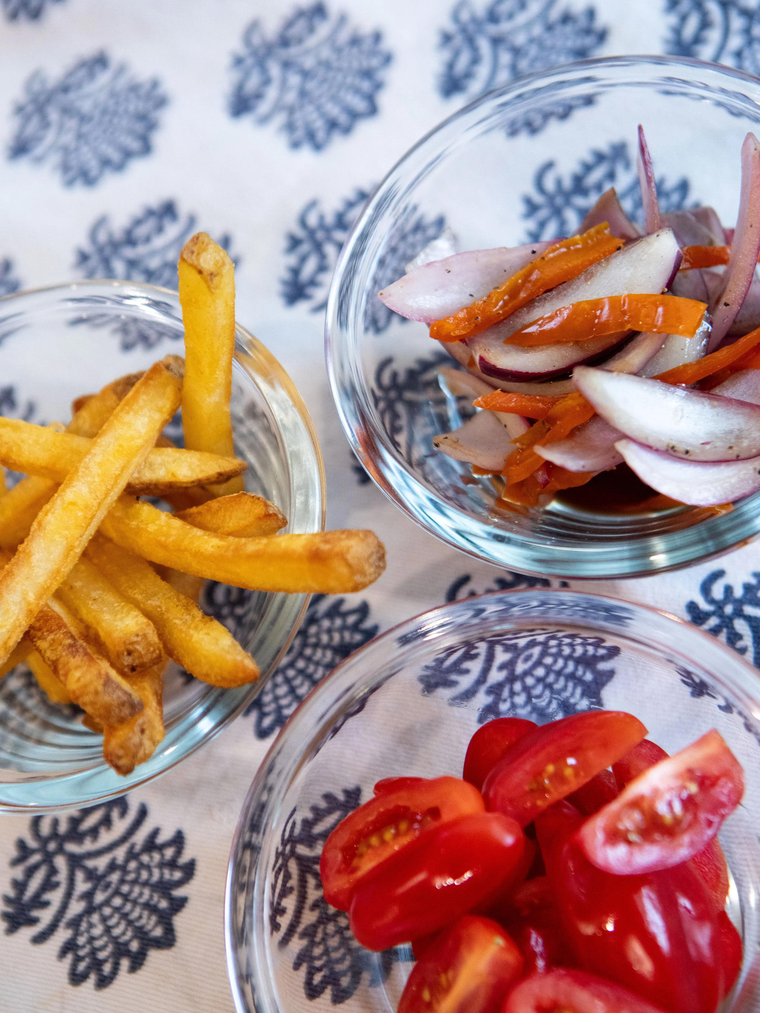 Bowls of French fries, pickled onions with carrots, and cherry tomatoes on a patterned tablecloth.
