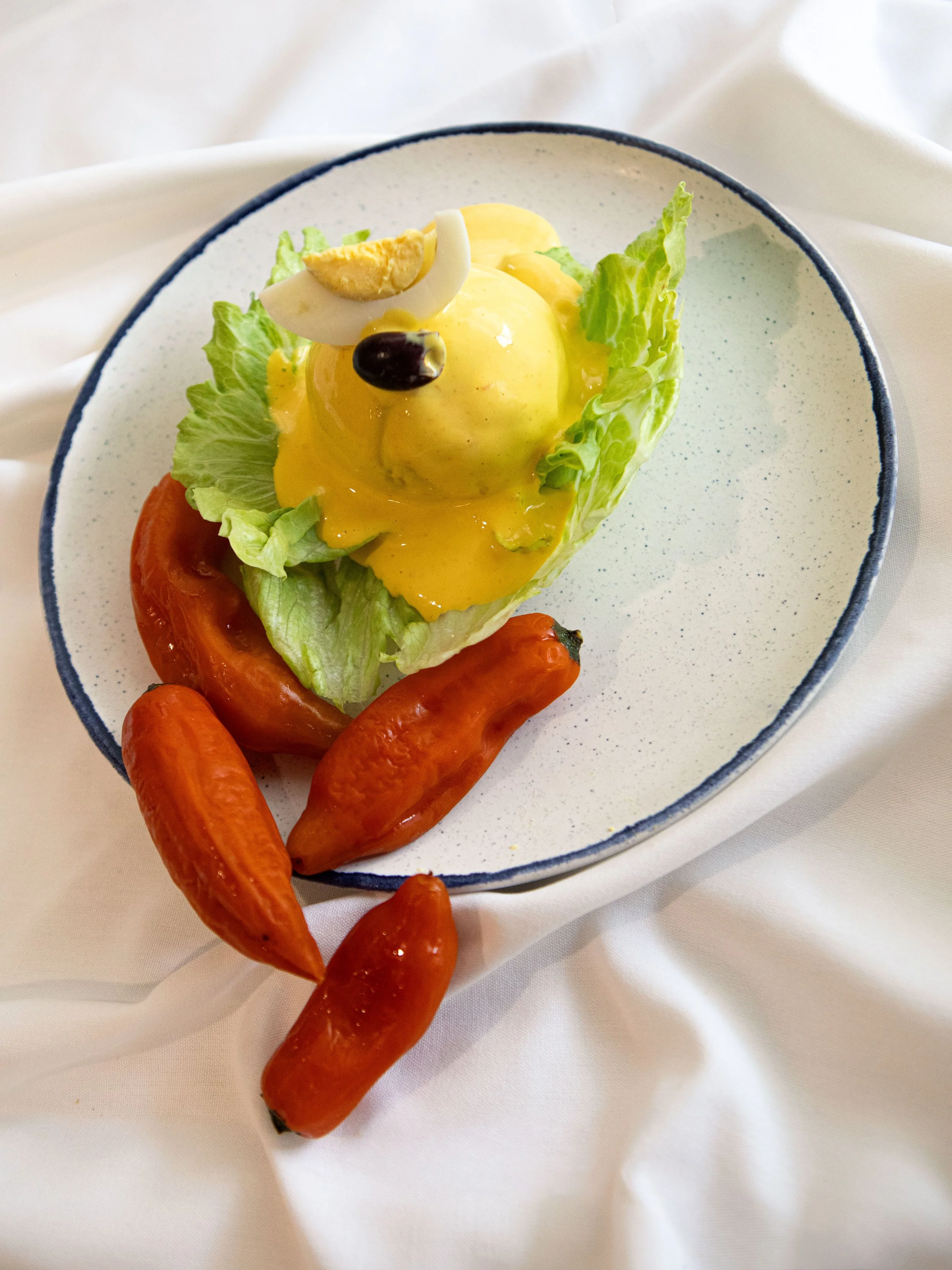 Deviled egg on a bed of lettuce in a white bowl, garnished with sliced hard-boiled egg and a black olive on top, served with small red hot chili peppers on the side.