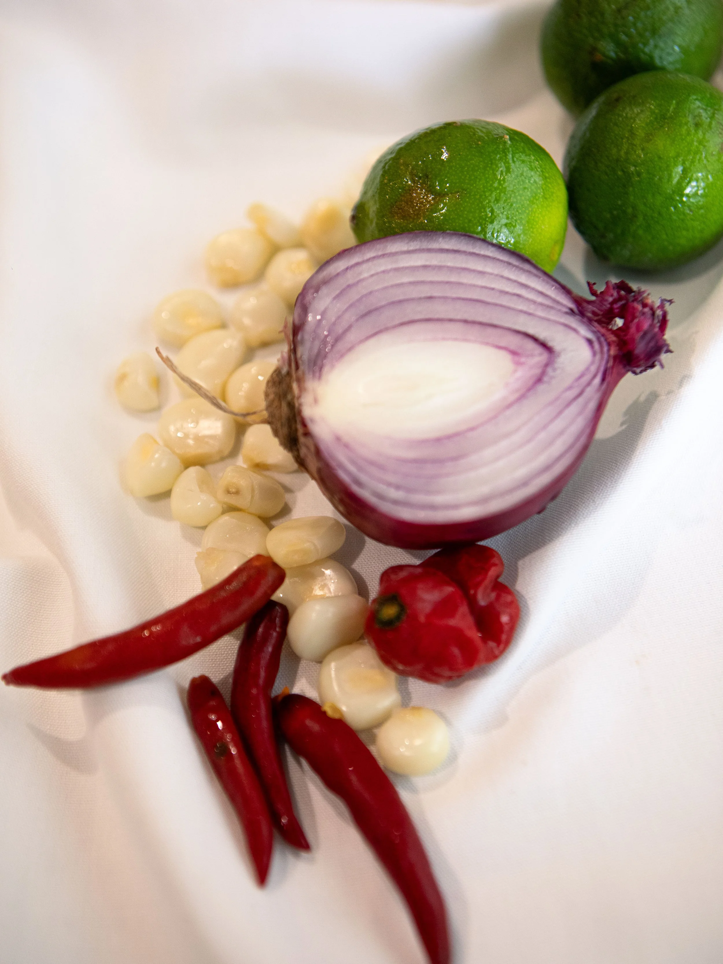 Limes, red chili peppers, chopped red onion, garlic cloves, and a red habanero pepper on a white surface.