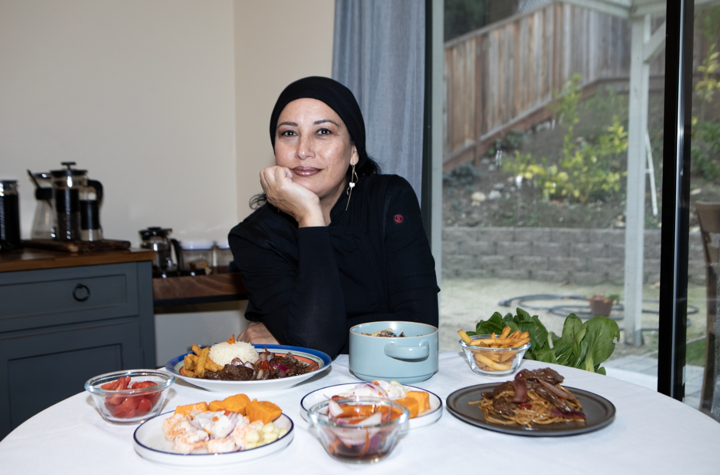 Woman in black hijab sitting at a table with various dishes, including rice, beef, pasta, salads, and a bowl of soup, in a kitchen near a large window with a wooden fence outside.