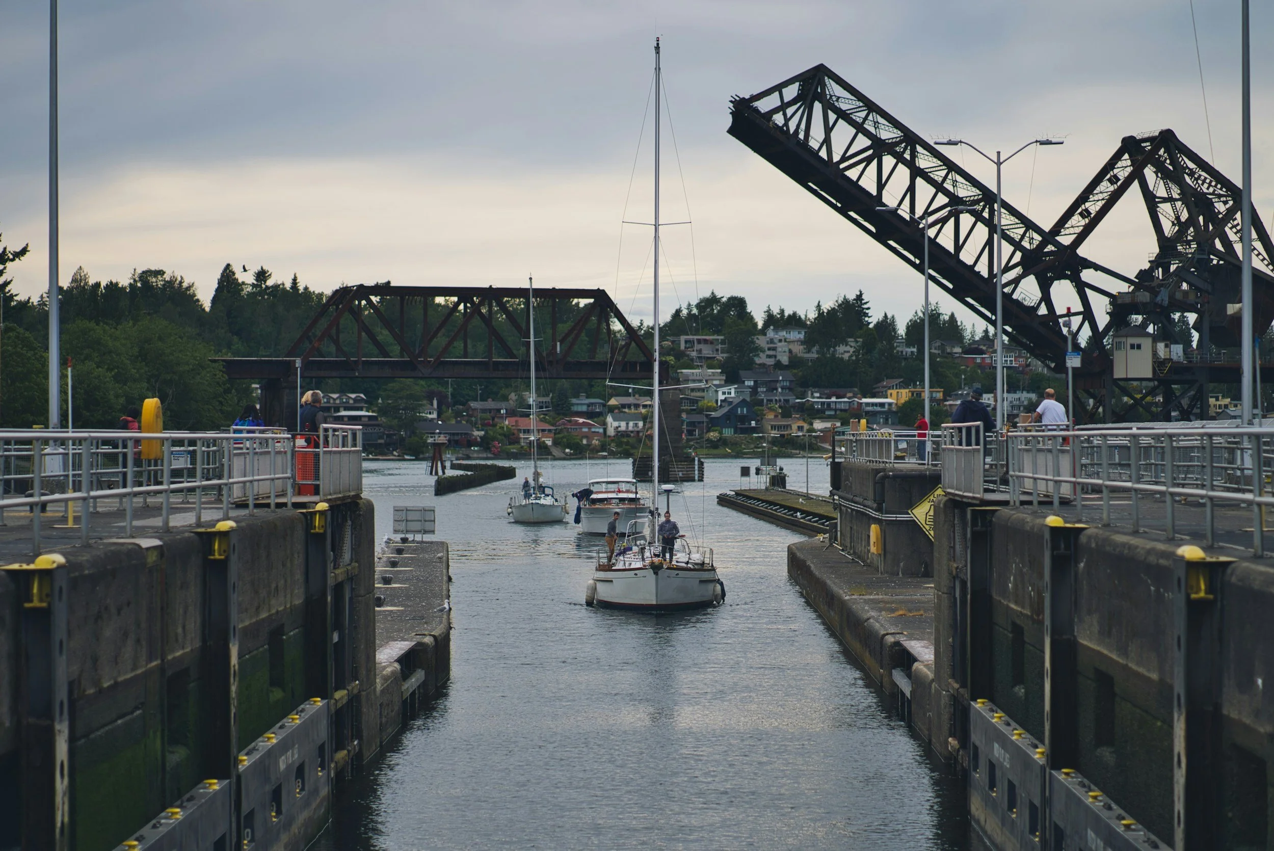 Boats passing through a canal lock with a raised lift bridge above, with houses and greenery in the background under a cloudy sky.