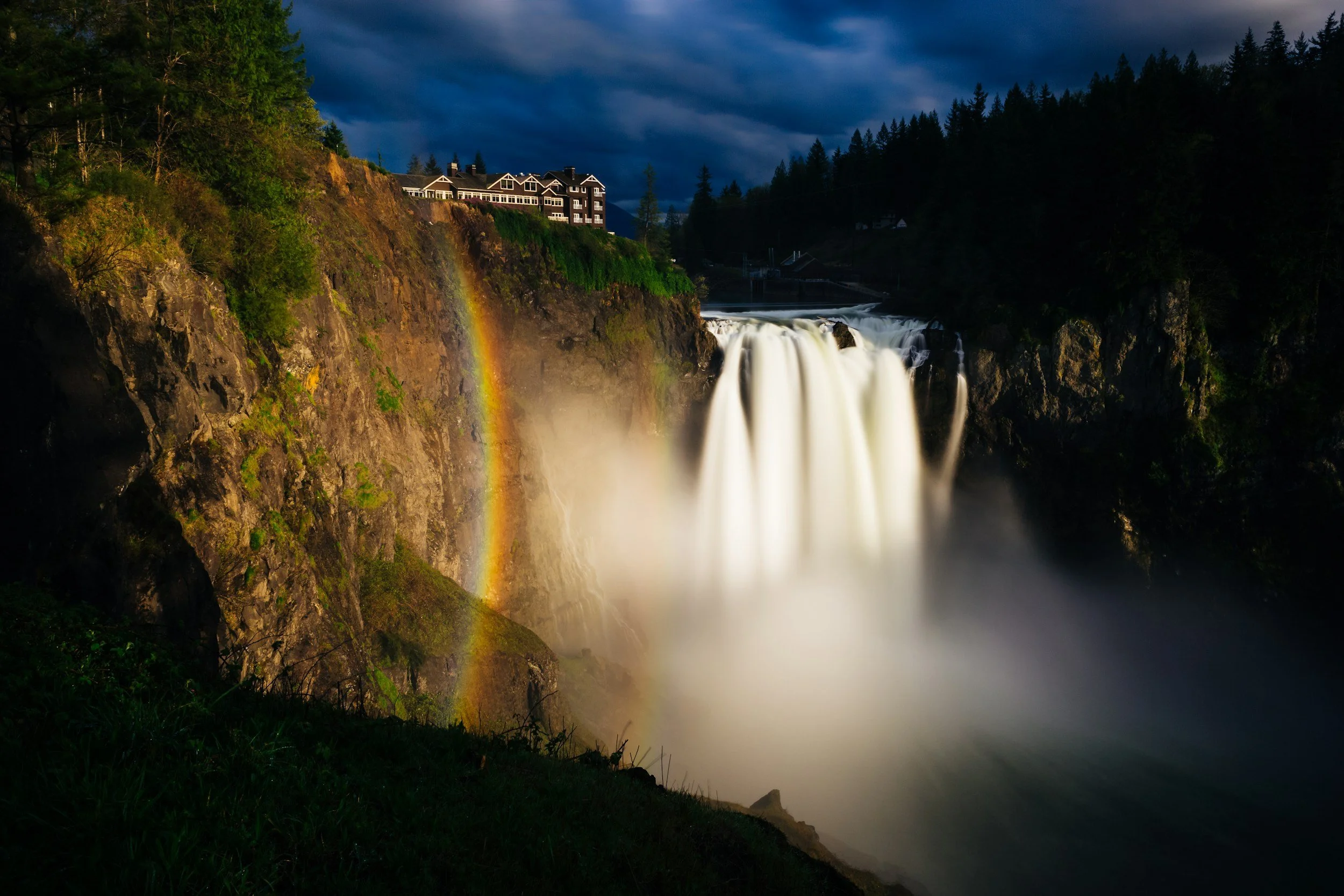 Waterfall at night with a rainbow, trees, a building on a cliff, and stormy sky.