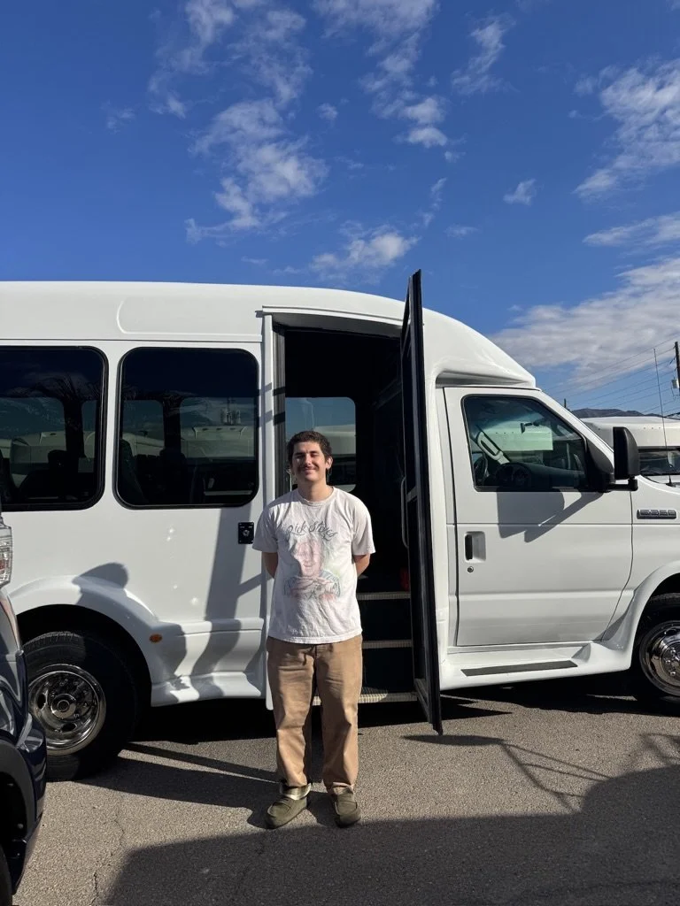A young man standing in front of a white bus with an open door, outdoors on a sunny day with a blue sky and scattered clouds.