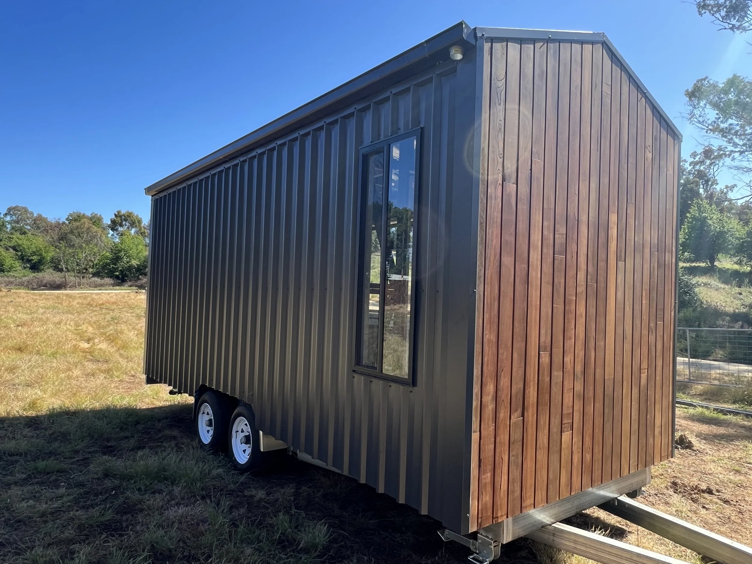 A modern tiny house trailer with a black metal exterior and wooden accent wall, set on a grassy field under a clear blue sky.