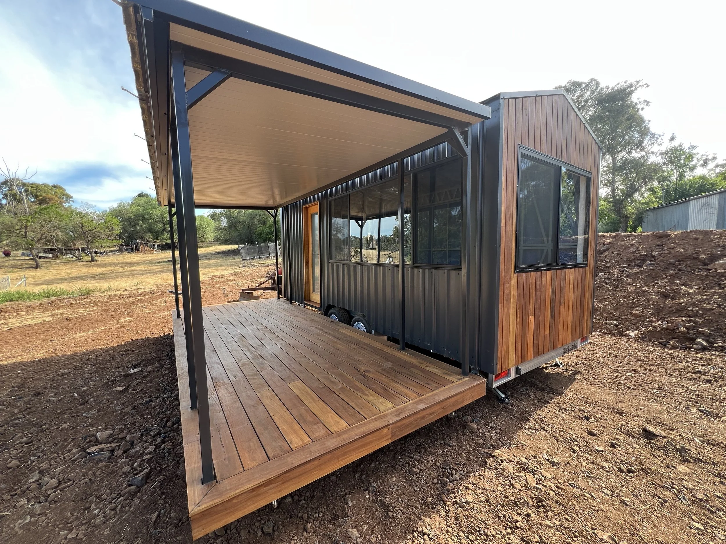 Mobile tiny house on a trailer with a wooden deck and an overhang, situated in a rural area with trees and dirt ground.
