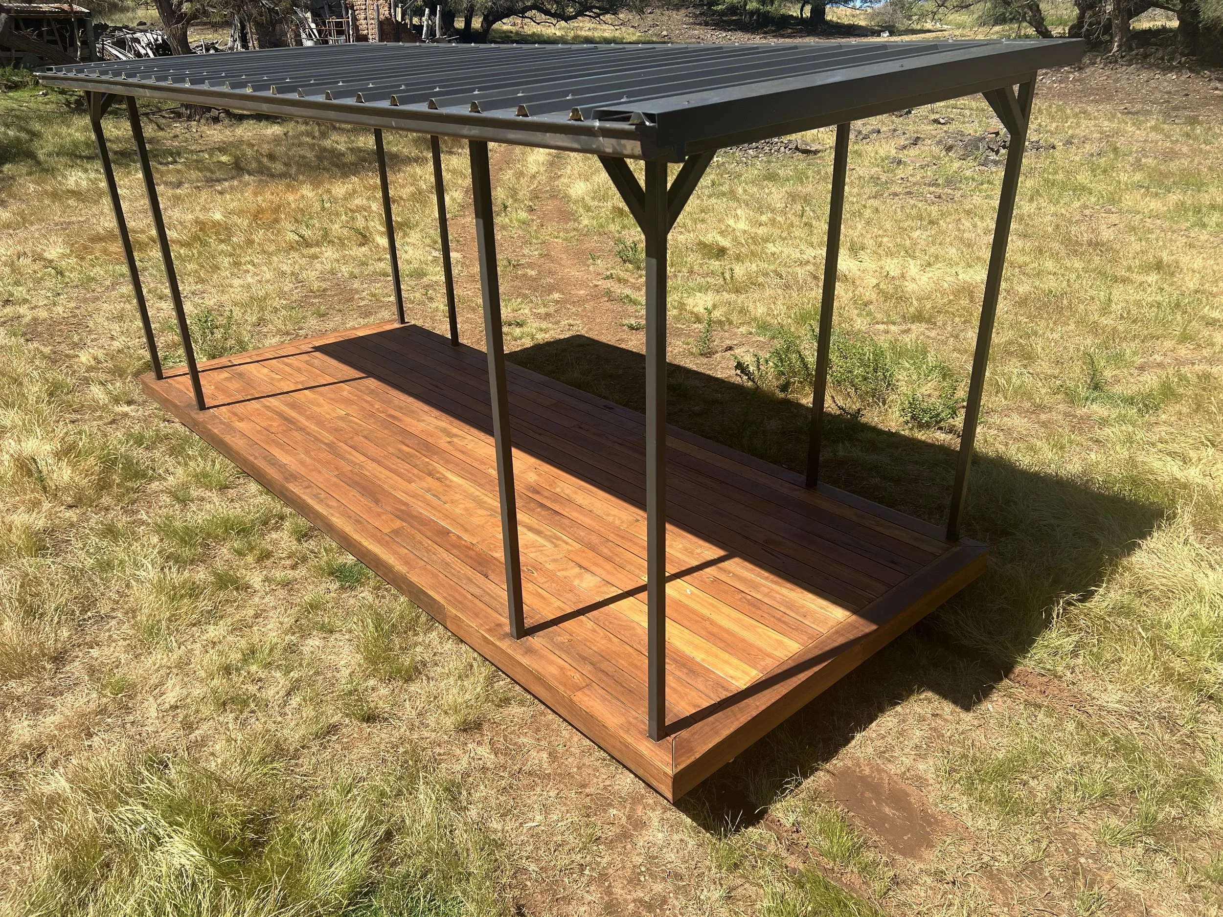 A small outdoor structure with a black metal roof, metal support beams, and a wooden floor, situated on a grassy area.