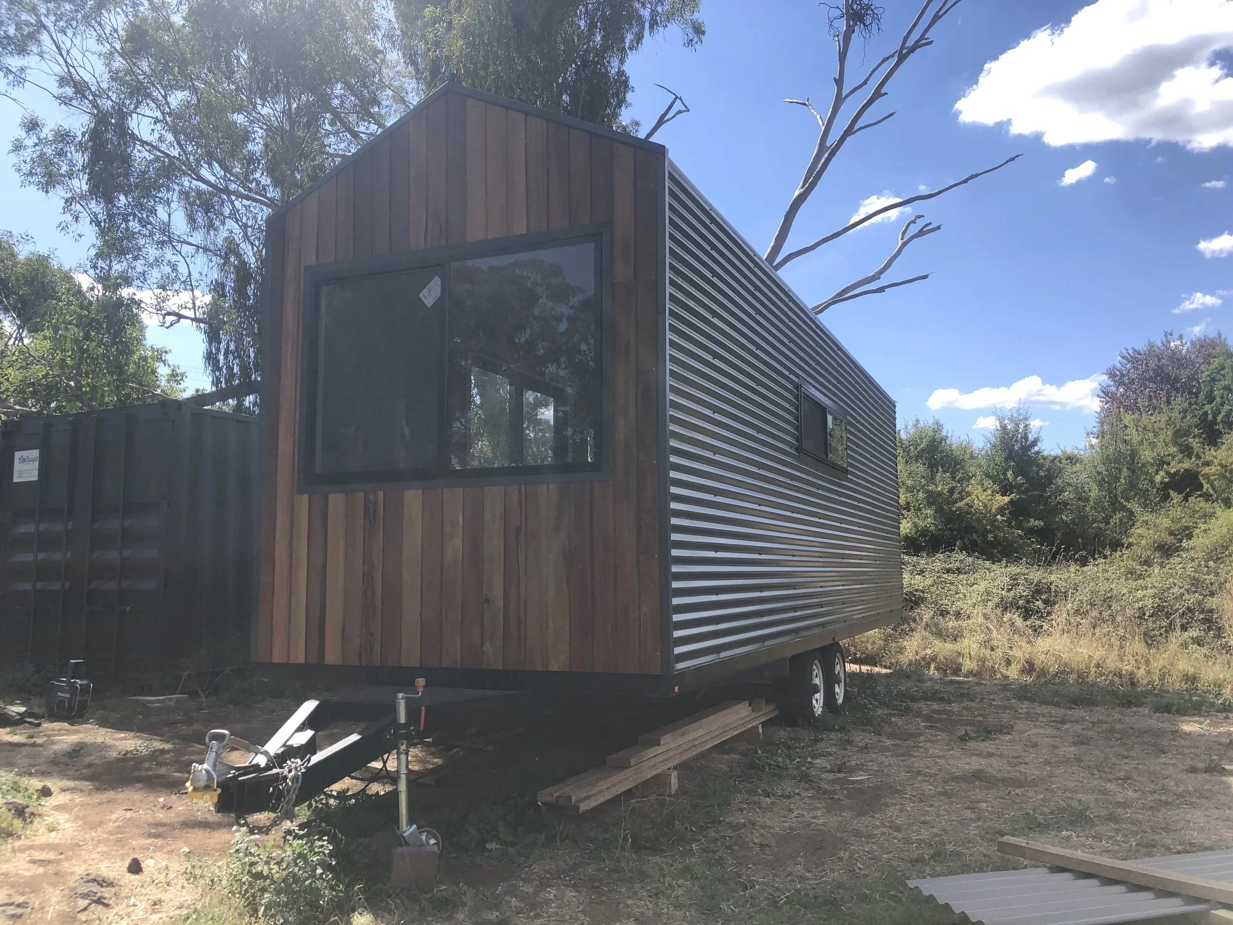 A tiny house on wheels with a metal siding exterior and large front window, set in a natural outdoor environment with trees and blue sky.