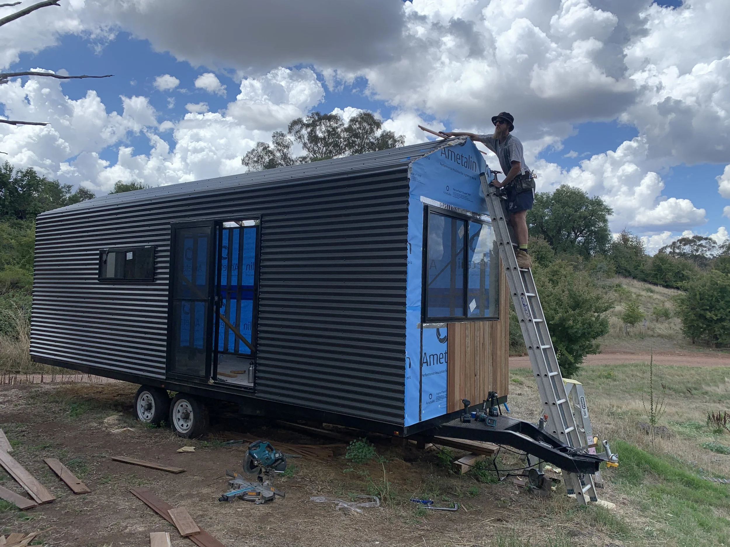 A person standing on a ladder working on the roof of a tiny house on wheels, with blue sky and clouds in the background.