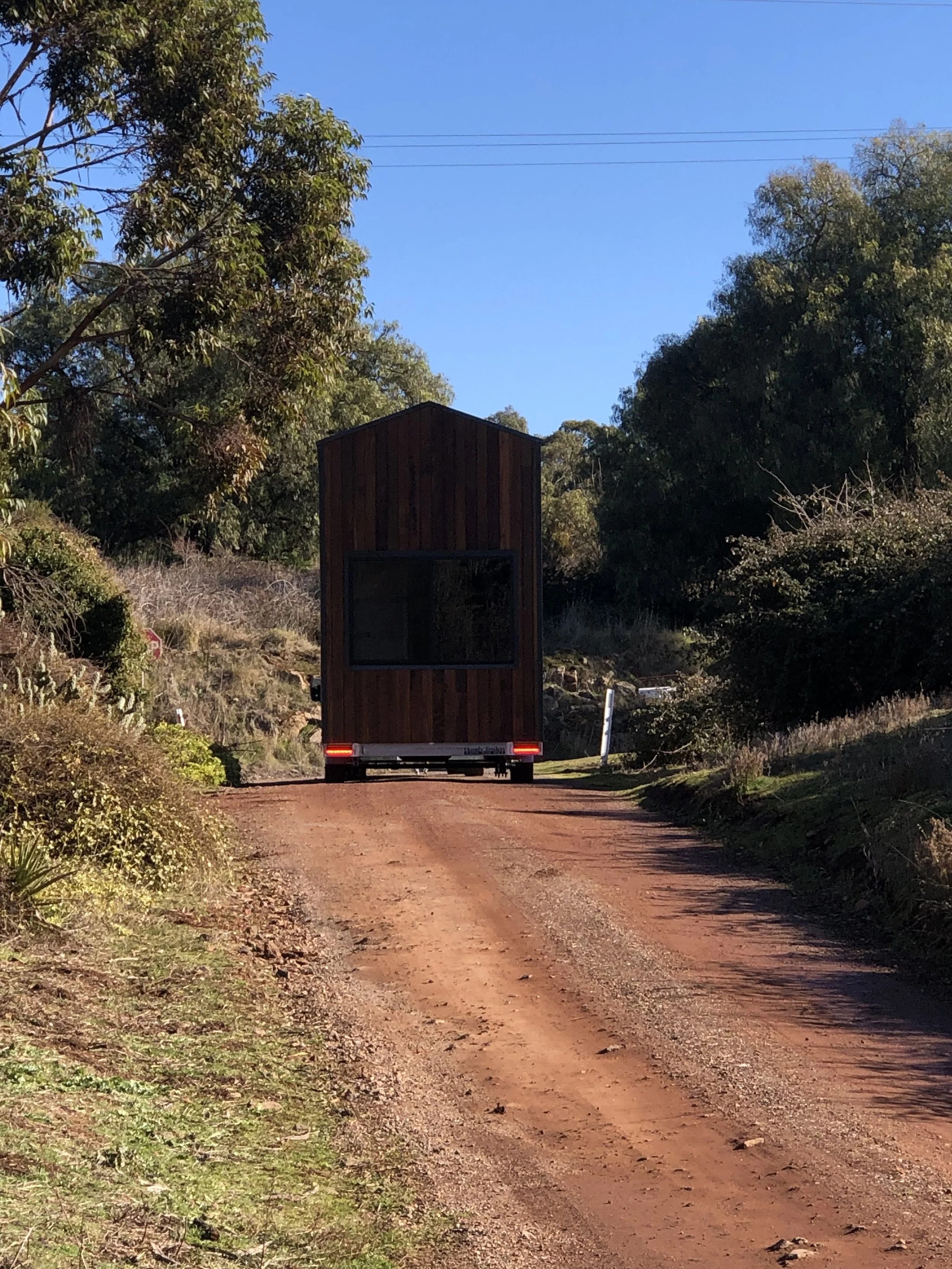 A tiny house on wheels with a wooden exterior is traveling along a dirt road surrounded by trees and vegetation, under a clear blue sky.