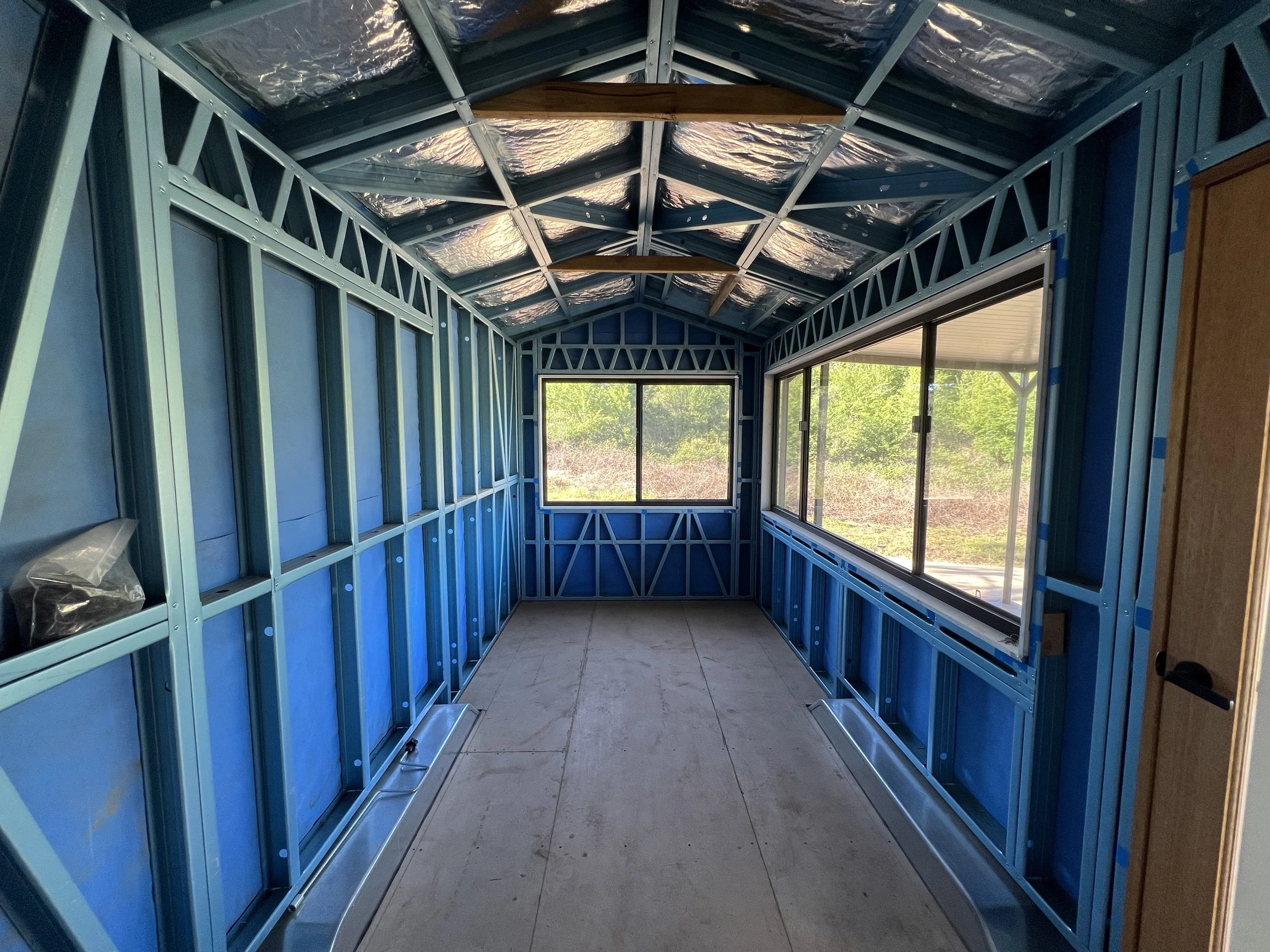 Interior view of a room under construction with exposed blue metal framing, drywall sheathing, large windows on two walls, and a concrete floor.