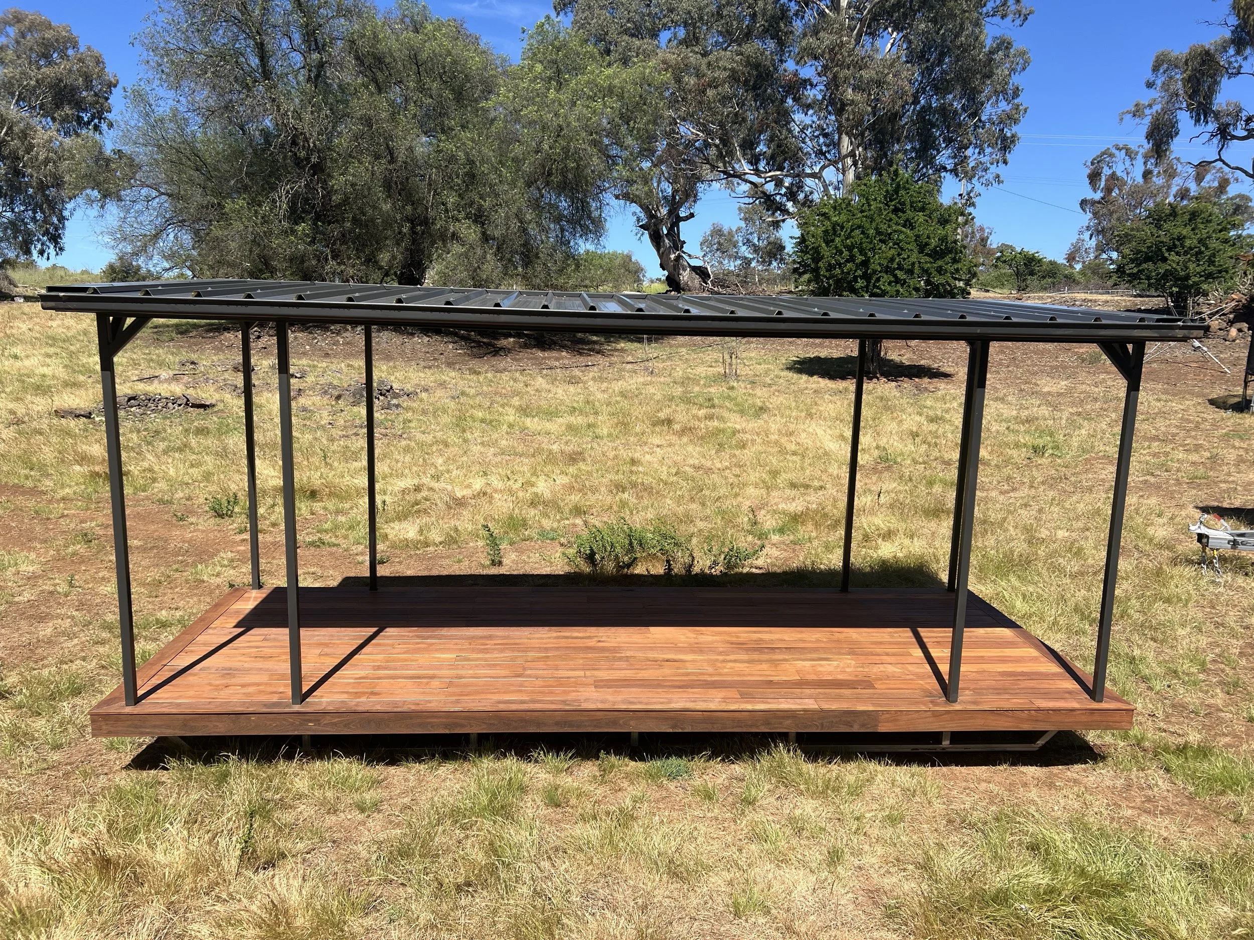 A black metal and wood deck with a metal roof outdoors on grass, with trees and a clear blue sky in the background.