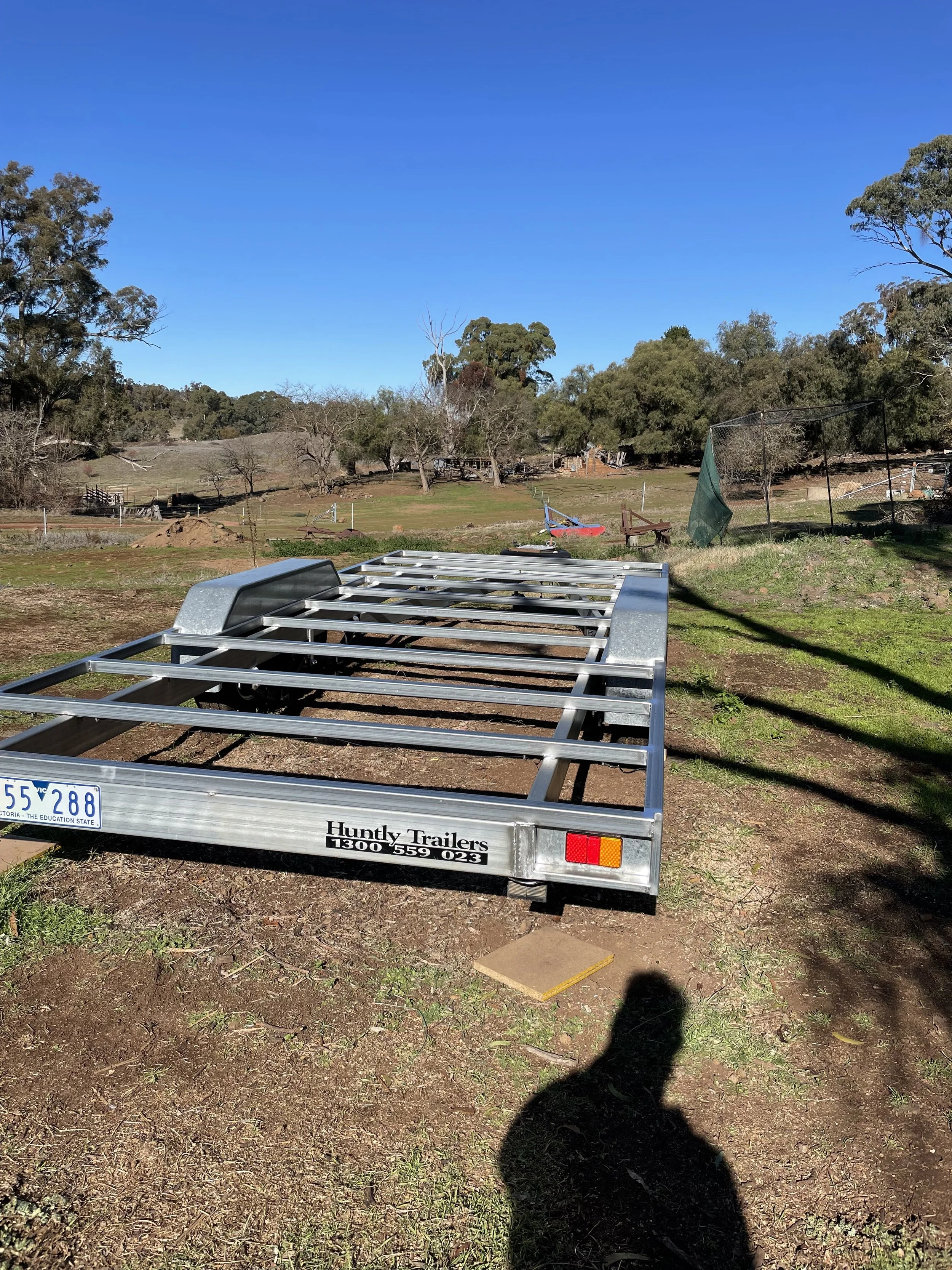 Empty metal trailer parked on dirt ground in an outdoor rural area, with trees and grass, blue sky, and a shadow of a person taking the photo.