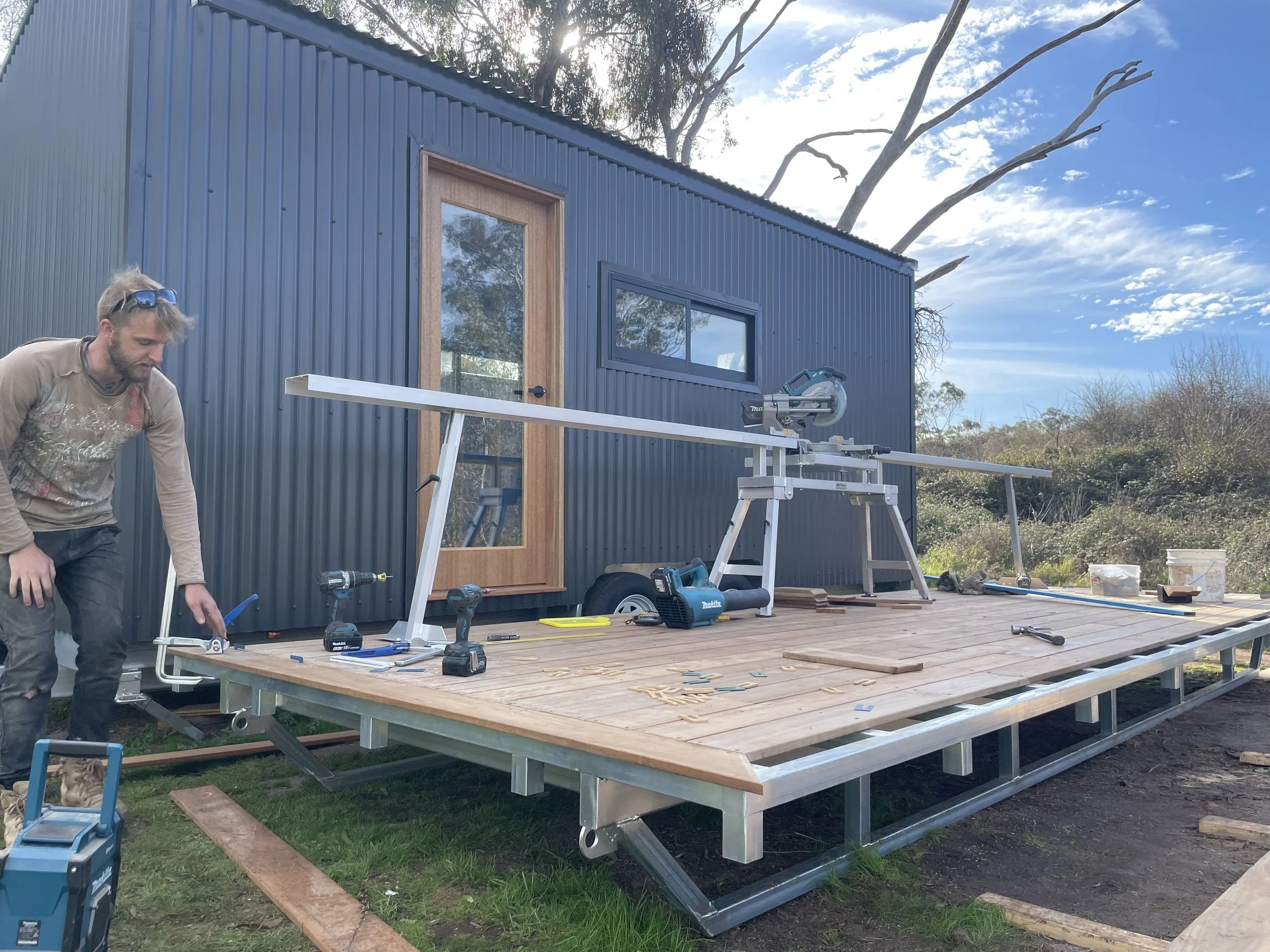 A man working on a small elevated deck in front of a tiny house, using a saw on a long wooden beam during daytime with a clear sky and trees in the background.