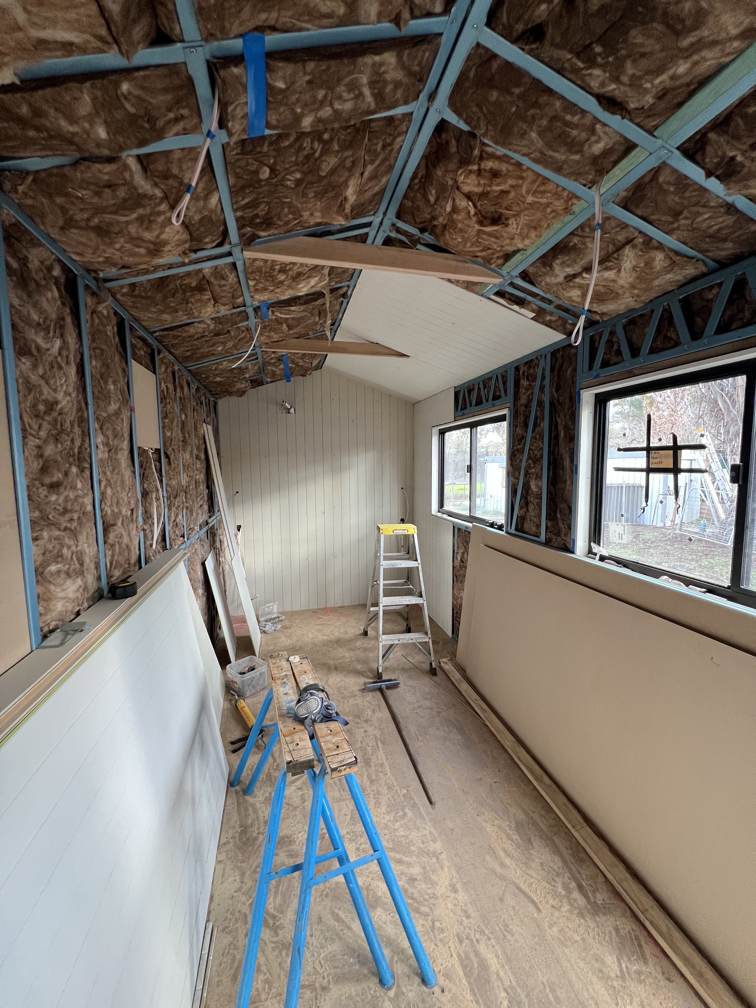 Room under construction with exposed insulation, metal framing, and partially installed interior wall panels. Construction tools and materials are scattered around, including a step ladder, saw horses, and drywall sheets.