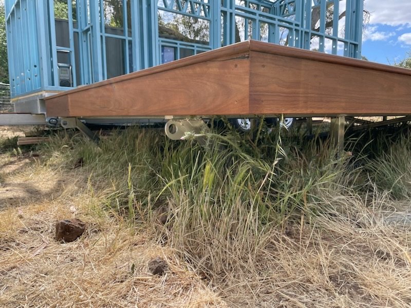 Close-up of a house under construction with a blue framing structure and a wooden foundation, supported by a trailer, with grassy ground beneath.