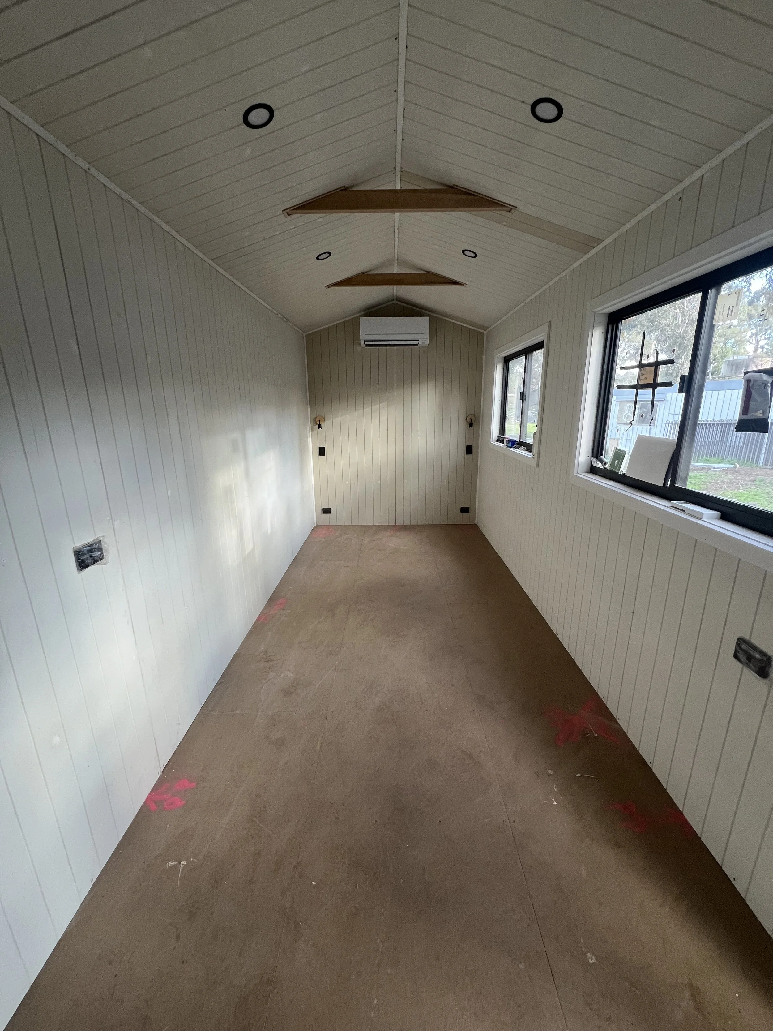 Empty room under construction with unfinished flooring, white paneled walls, ceiling with recessed lights, and two ceiling fans. One side has two windows showing an outdoor yard.